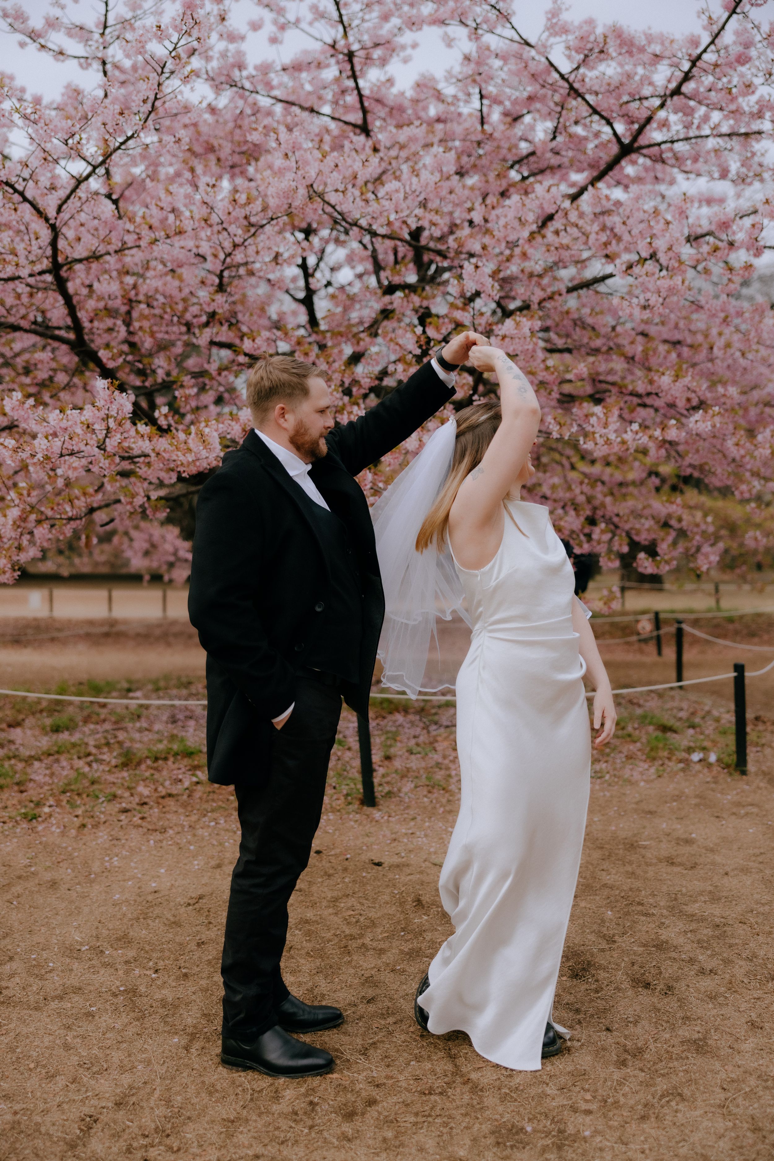 A couple dancing under pink cherry blossoms with the bride in white dress and groom in black suit.