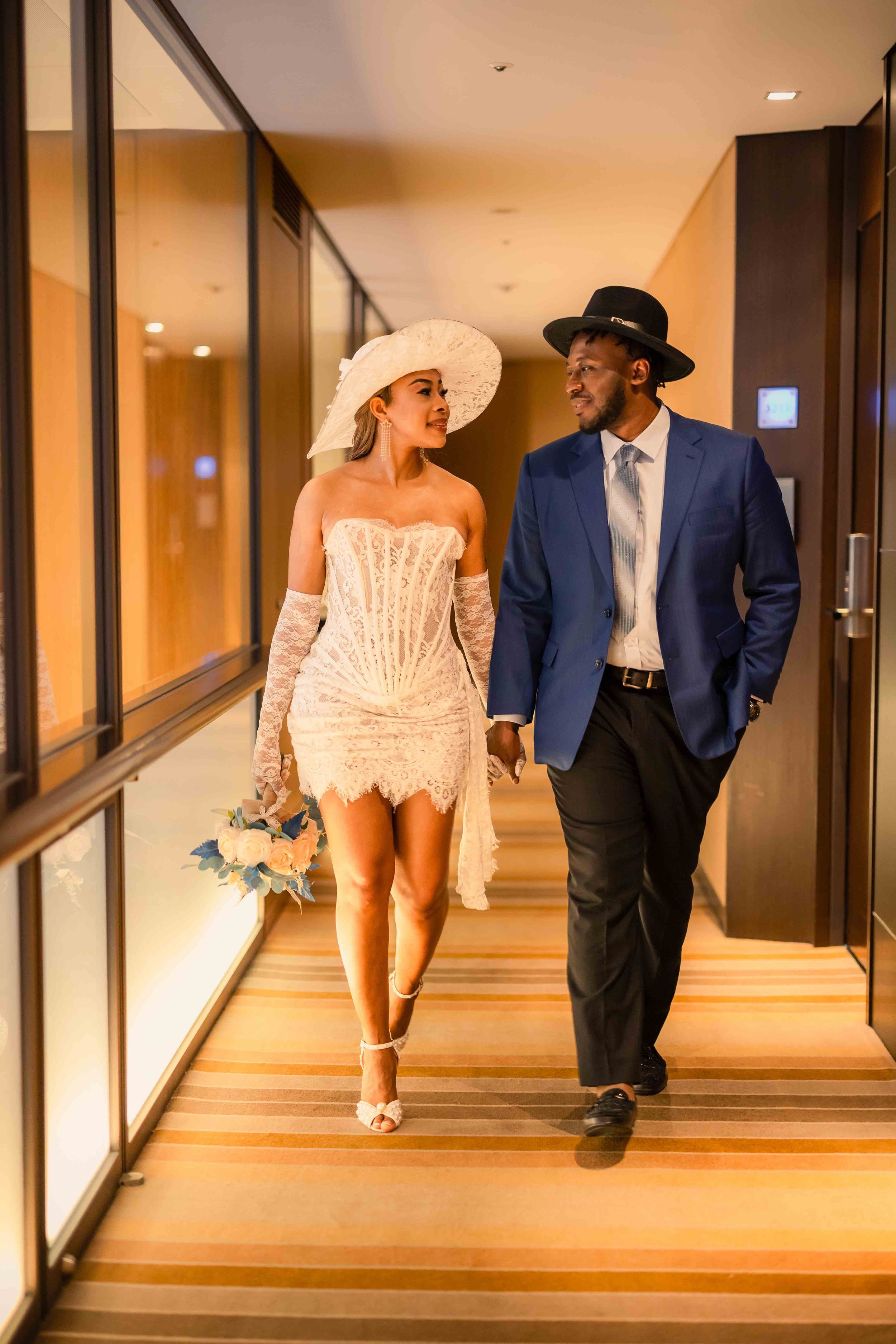 Stylish couple walking down hotel hallway, bride in short white dress and wide-brimmed hat.