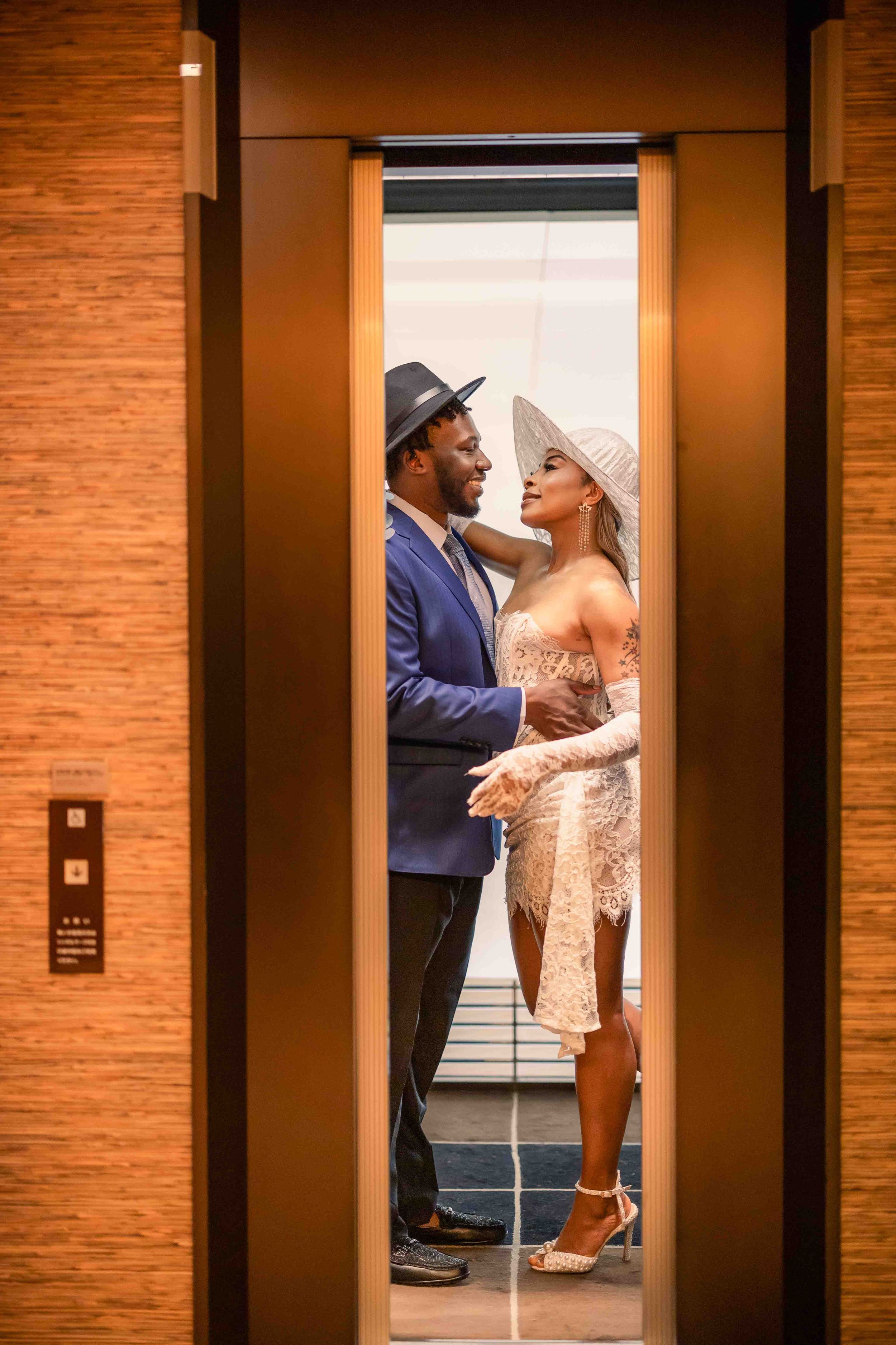 Romantic couple pose in elevator doorway wearing formal attire and matching white hats.