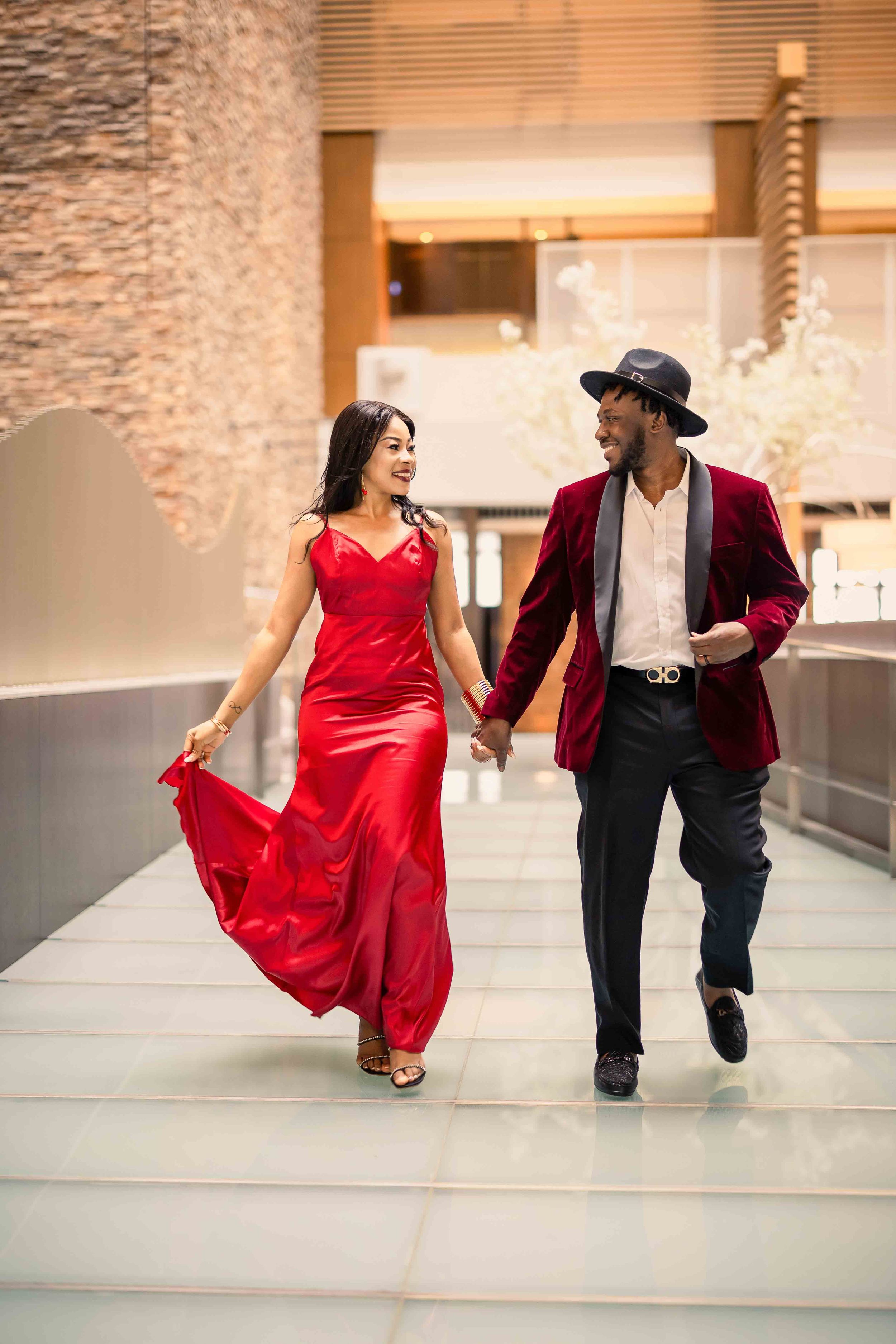 Stylish couple walking hand in hand, with flowing red gown and burgundy velvet blazer.