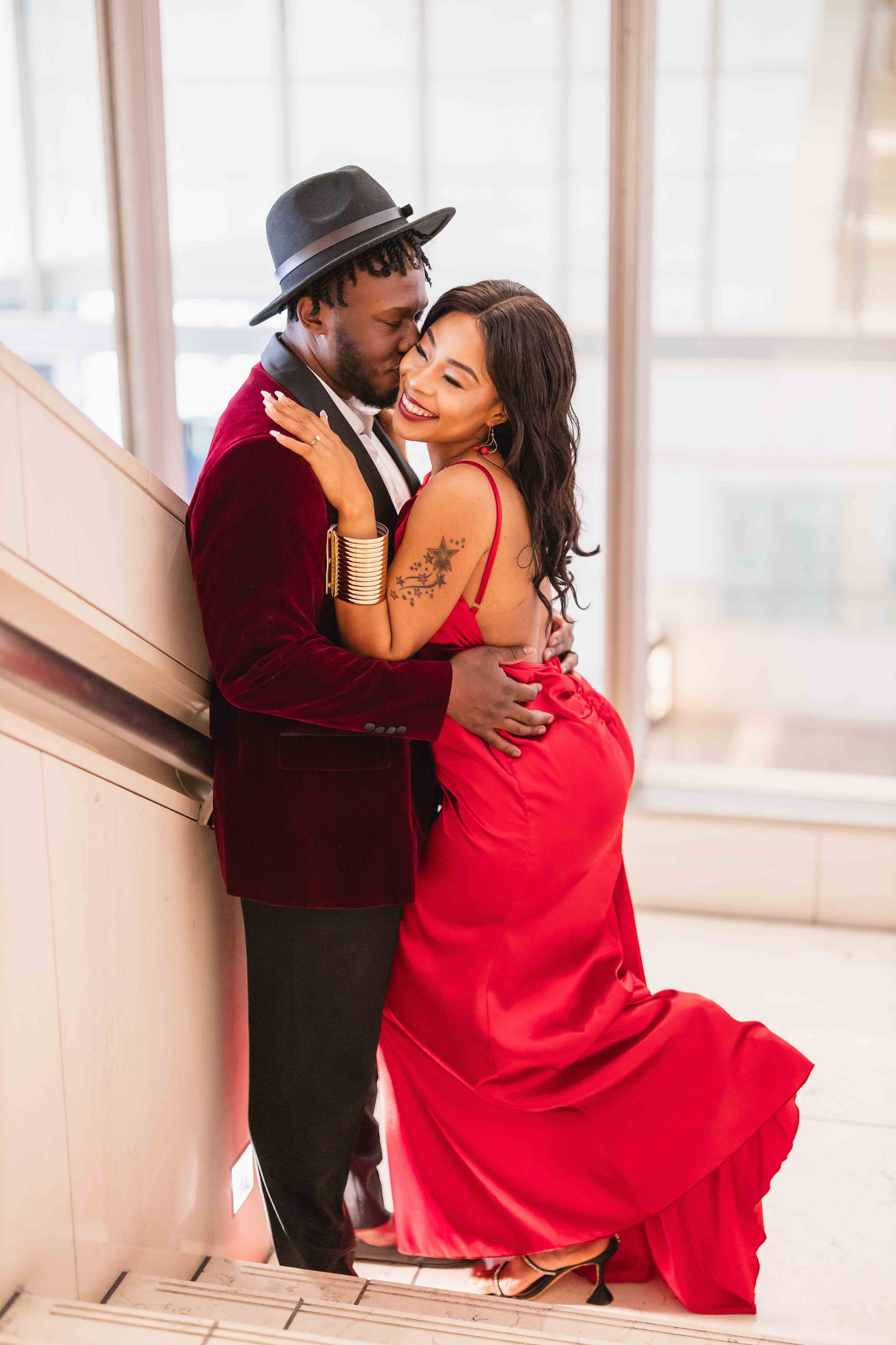Affectionate couple embrace on staircase wearing formal red and burgundy attire.