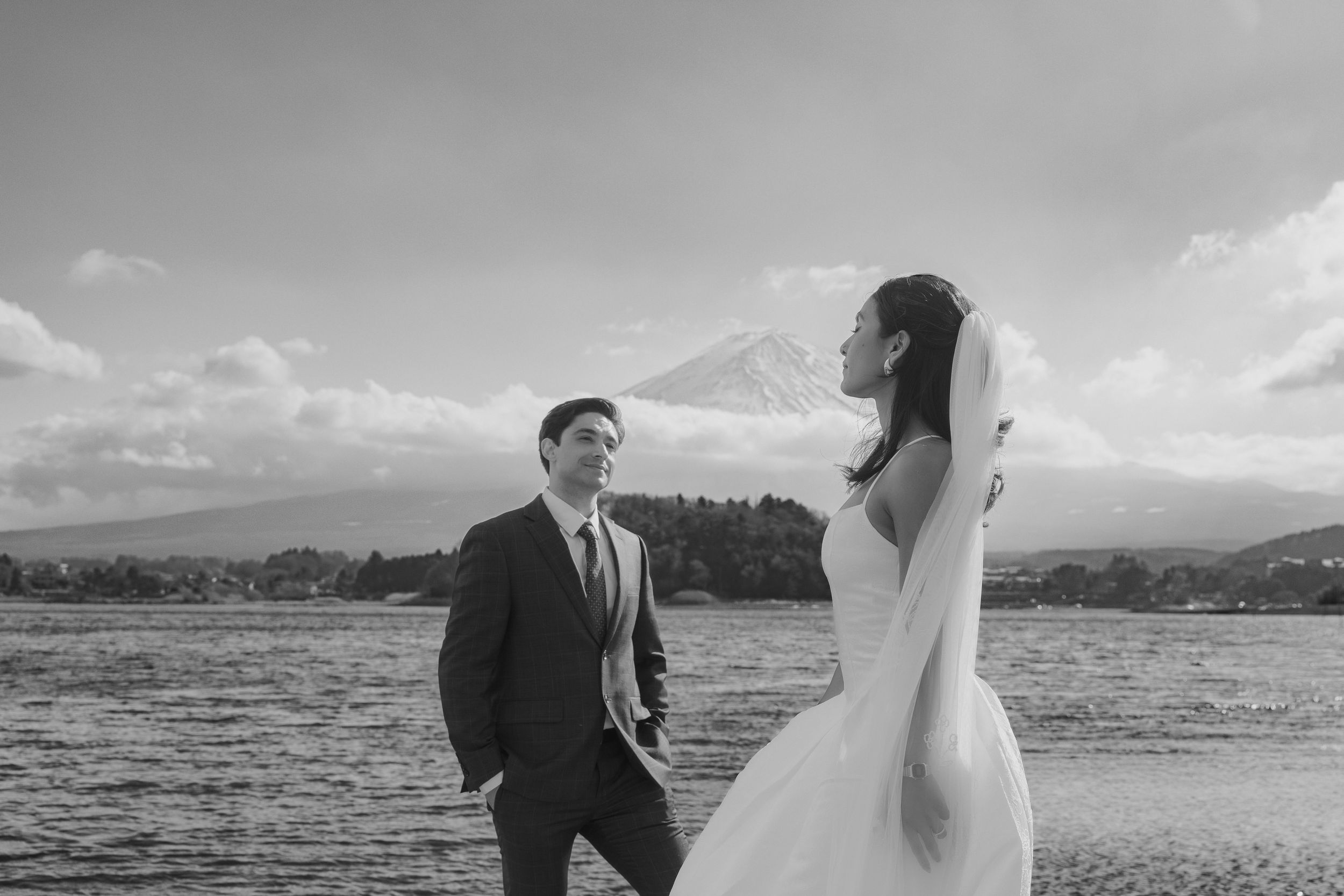 A happy couple shares a romantic moment on a beach with mountains in the background during their wedding photoshoot.