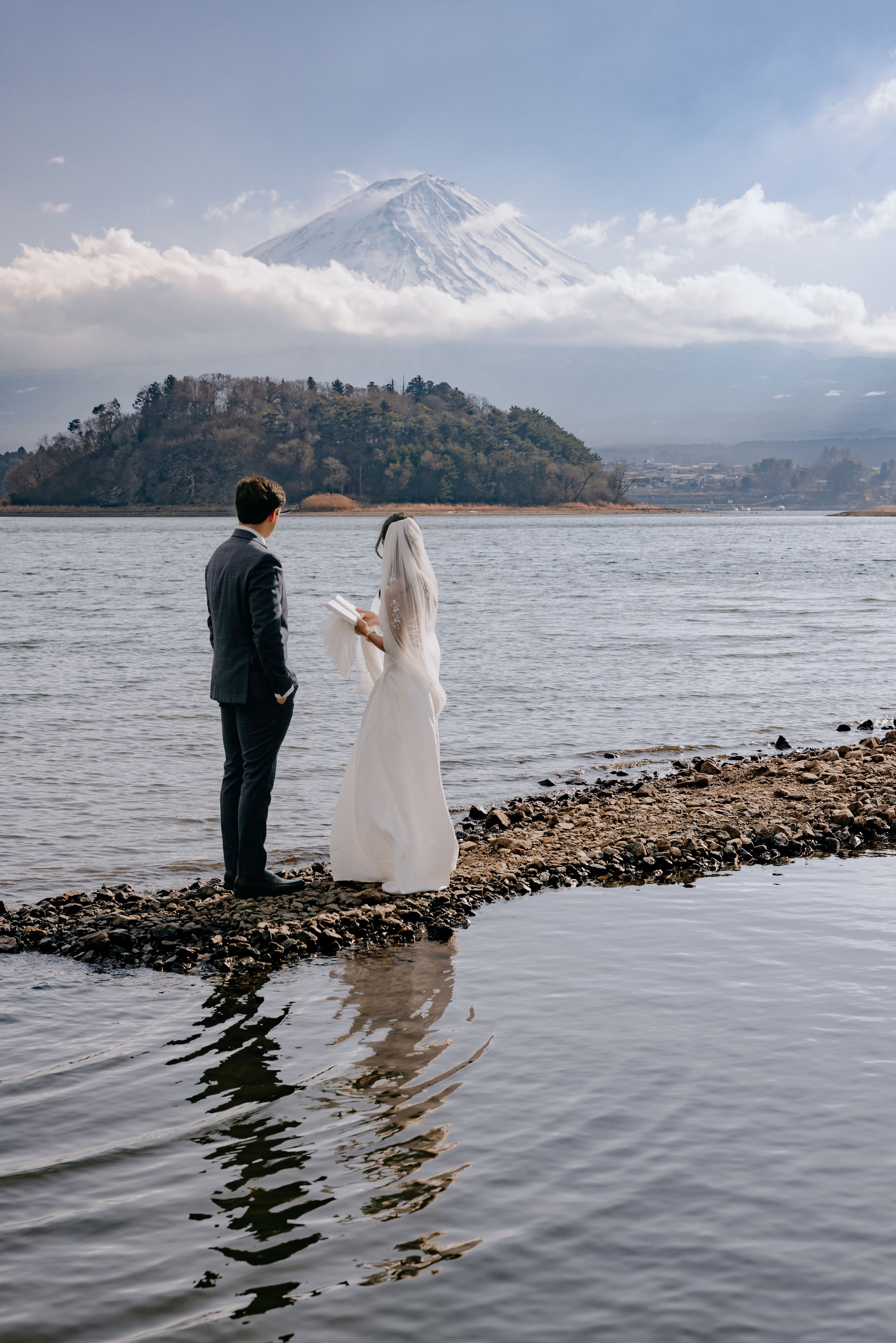 A couple in wedding attire stands at the edge of Lake Kawaguchiko with Mount Fuji rising majestically in the background.