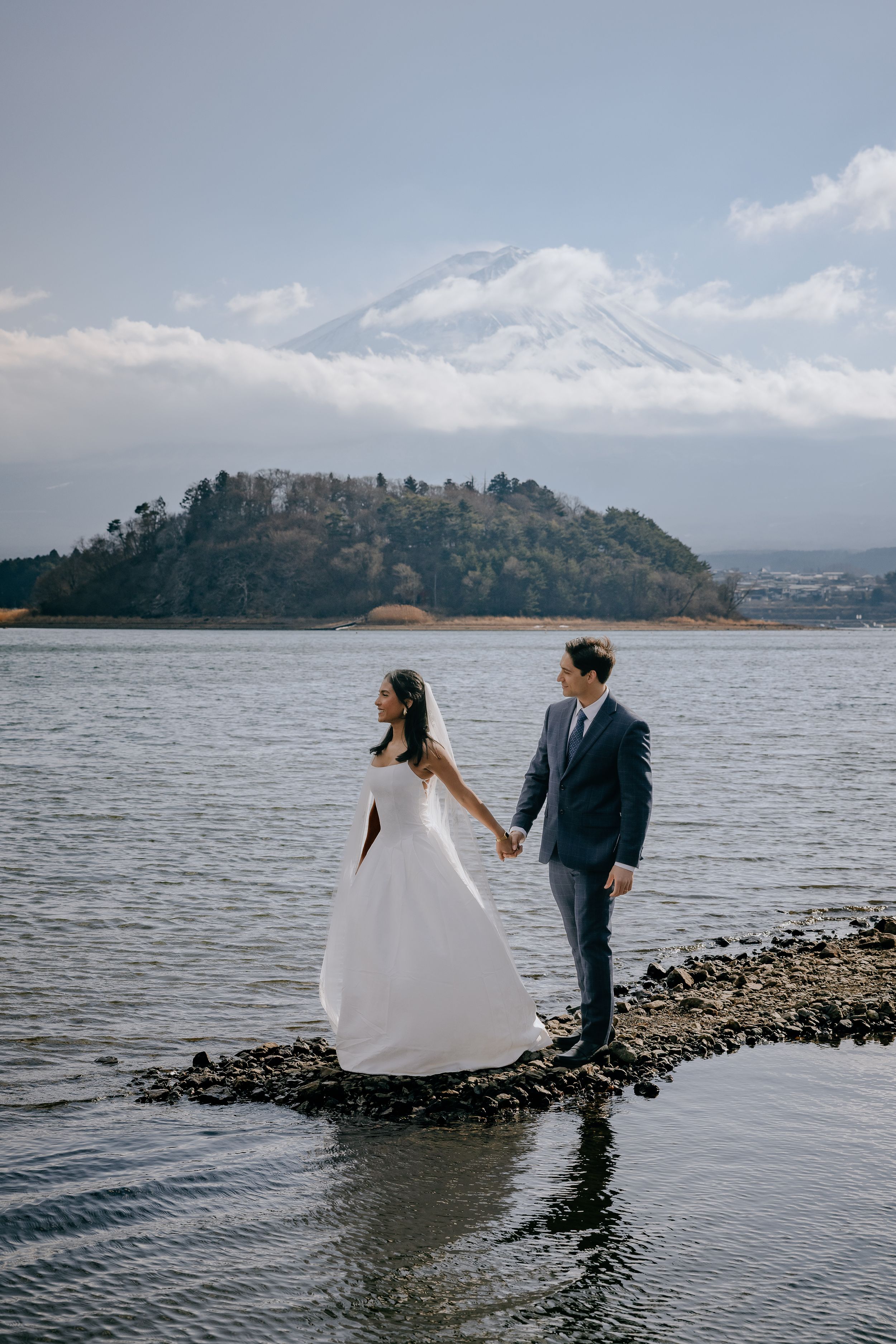 A couple stands along a rocky shoreline with mountains and water in the background on their wedding day.