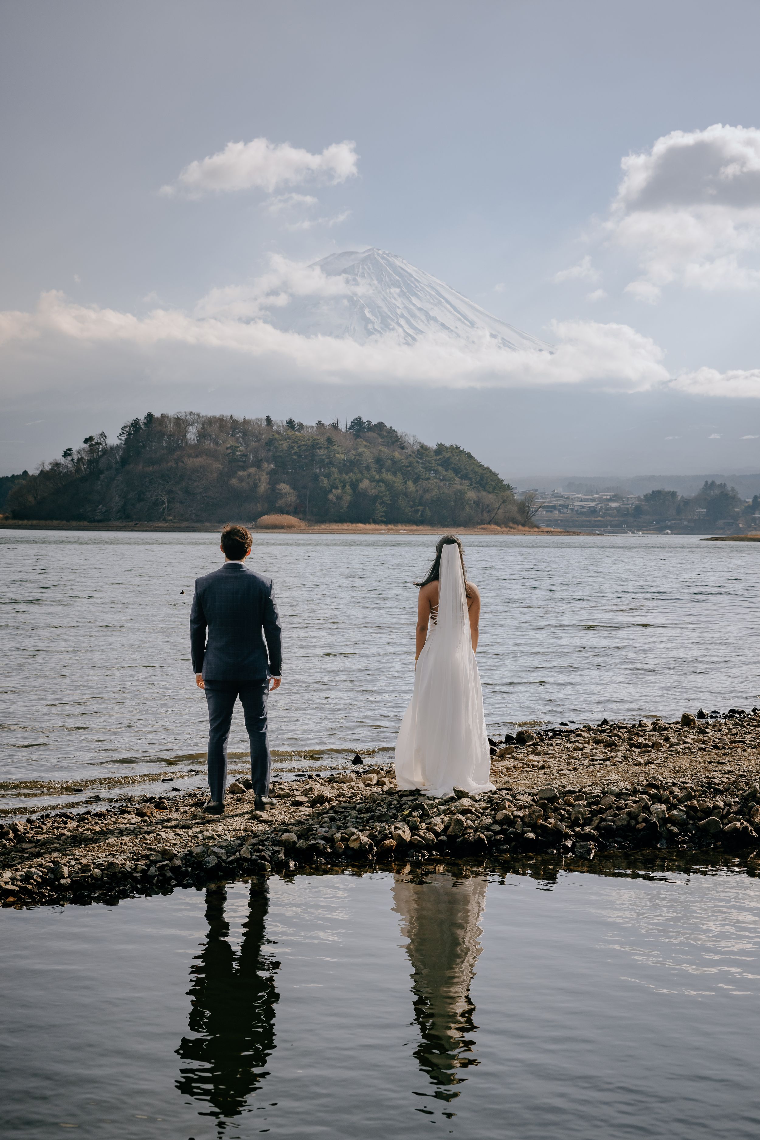 A couple in wedding attire stands at the edge of a rocky shoreline with mountains and water in the background.