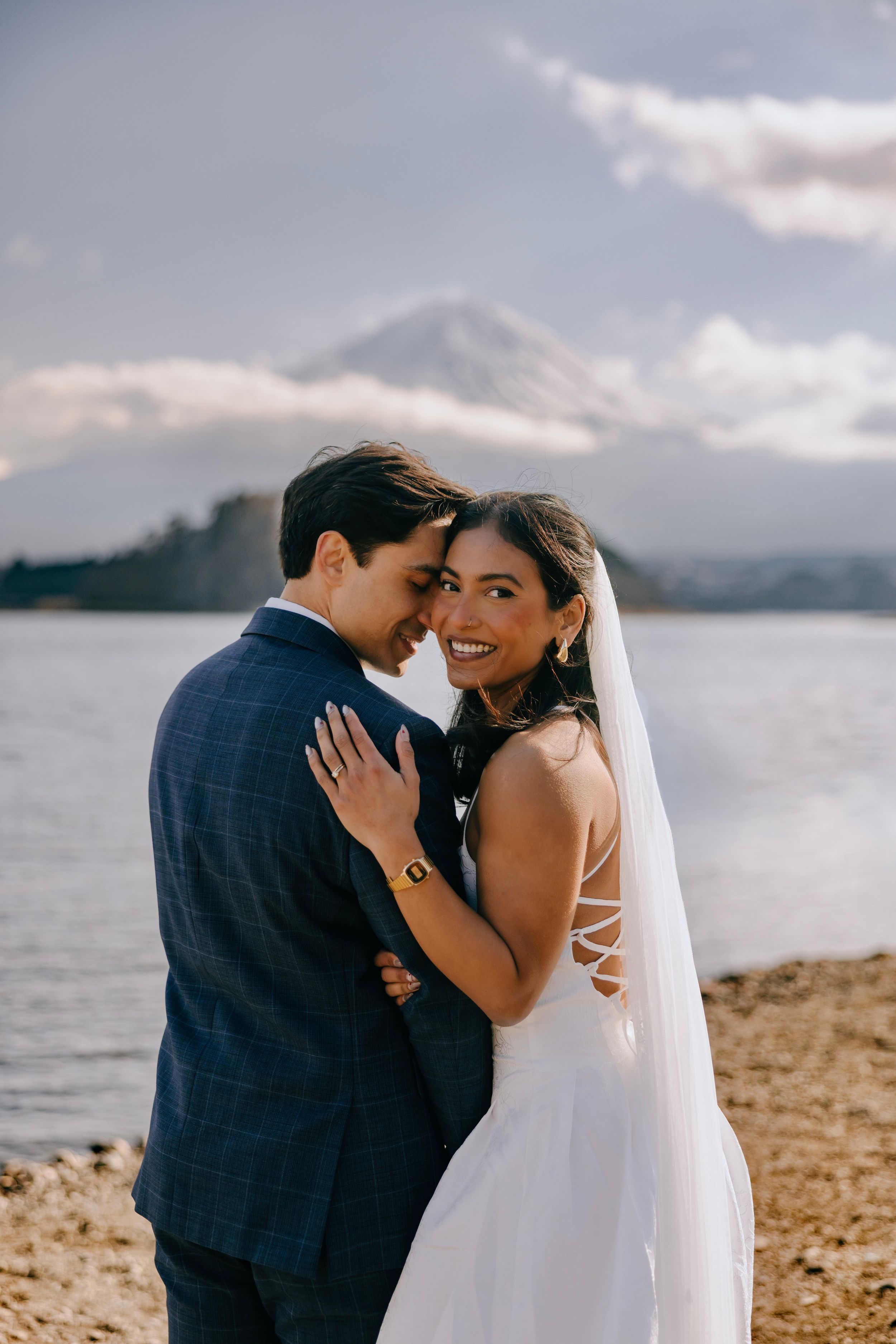 A couple shares intimate moments on a beach with mountains and water in the background during their wedding photoshoot.