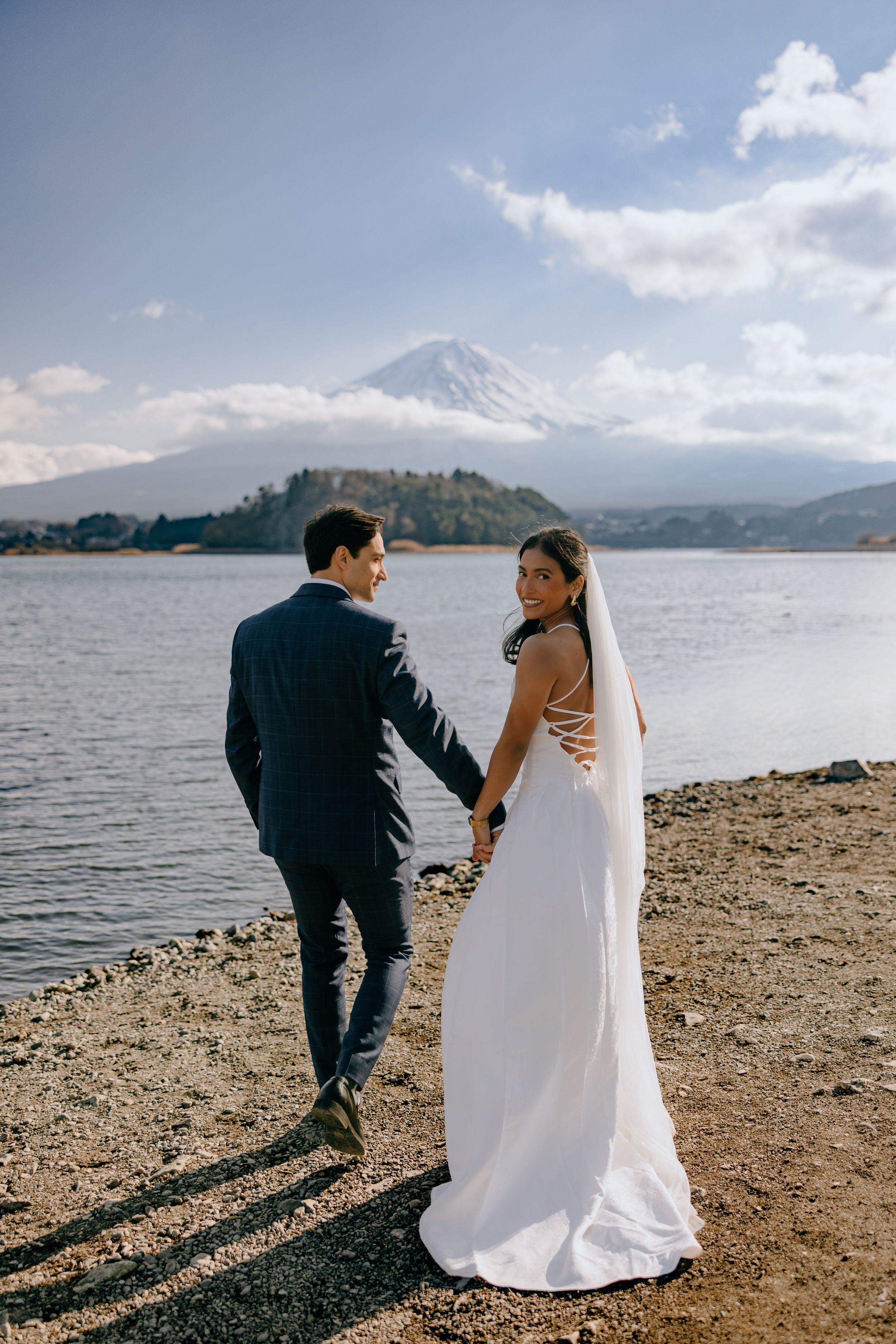 Couple in wedding attire walking along rocky shoreline with Mount Fuji and Lake Kawaguchiko in background.