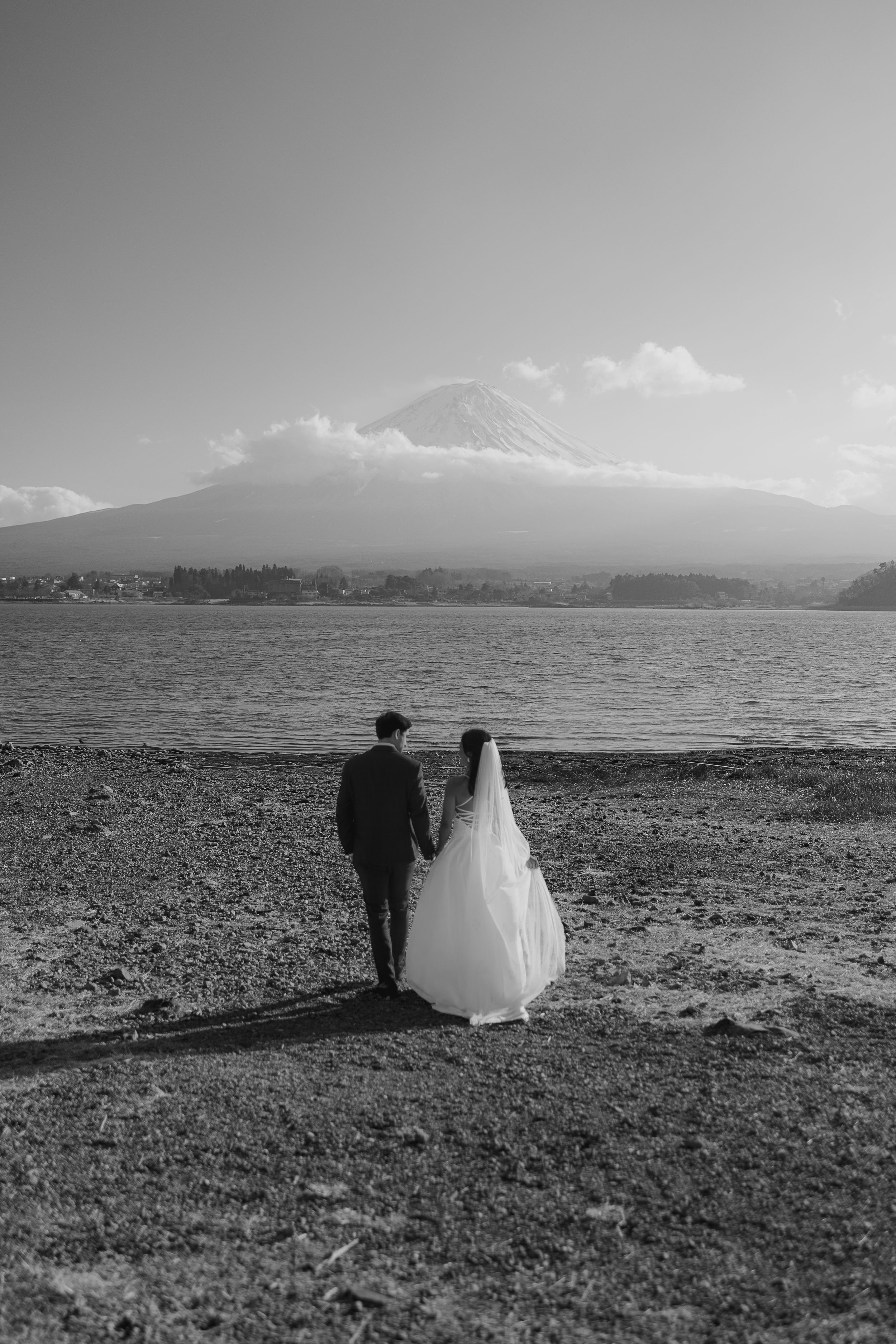 A bride and groom walk along a scenic lakeside meadow with mountains in the background in black and white.