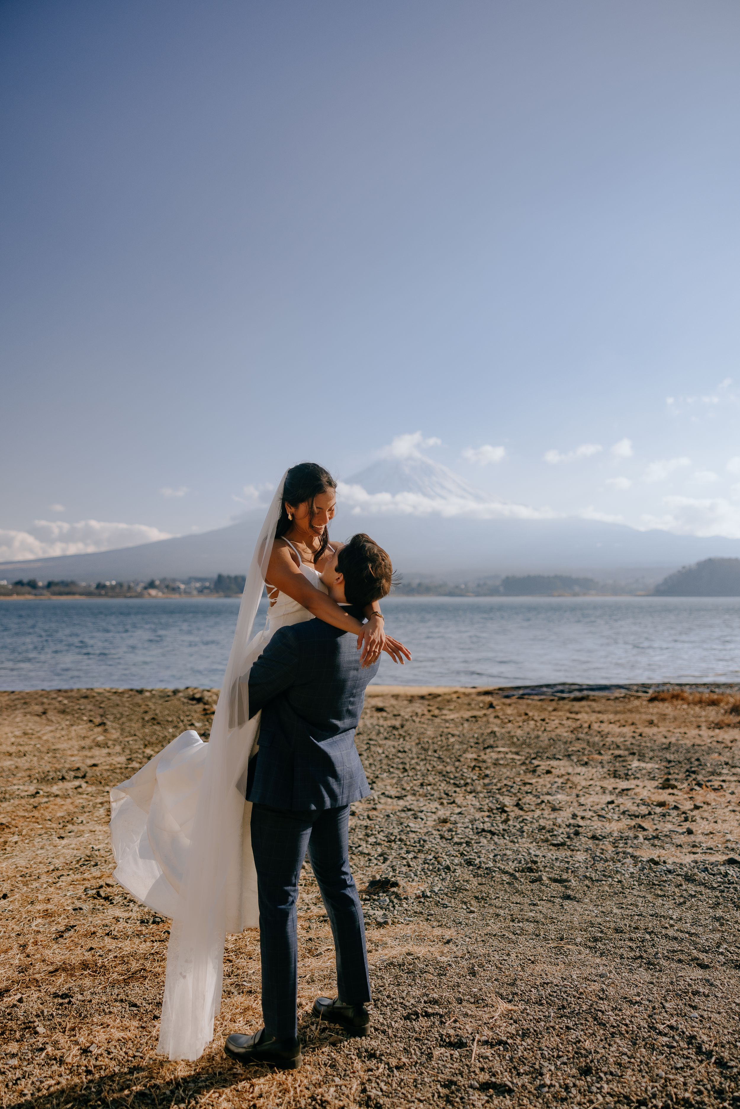 Couple embraces on a rocky beach shoreline with mountains and water in the background.