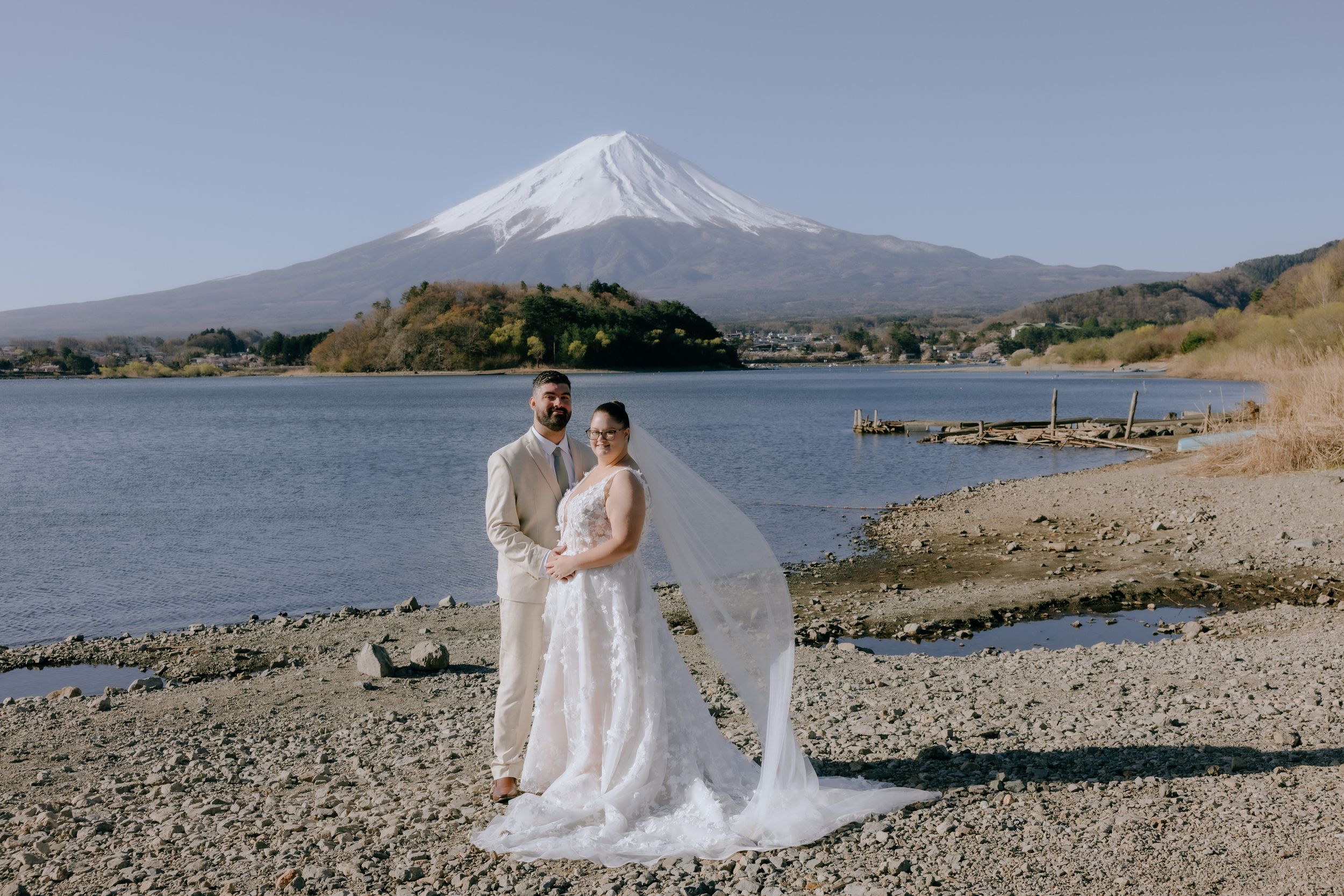 A wedding couple stands on the shore of Lake Kawaguchiko with snow-capped Mount Fuji rising majestically in the background.