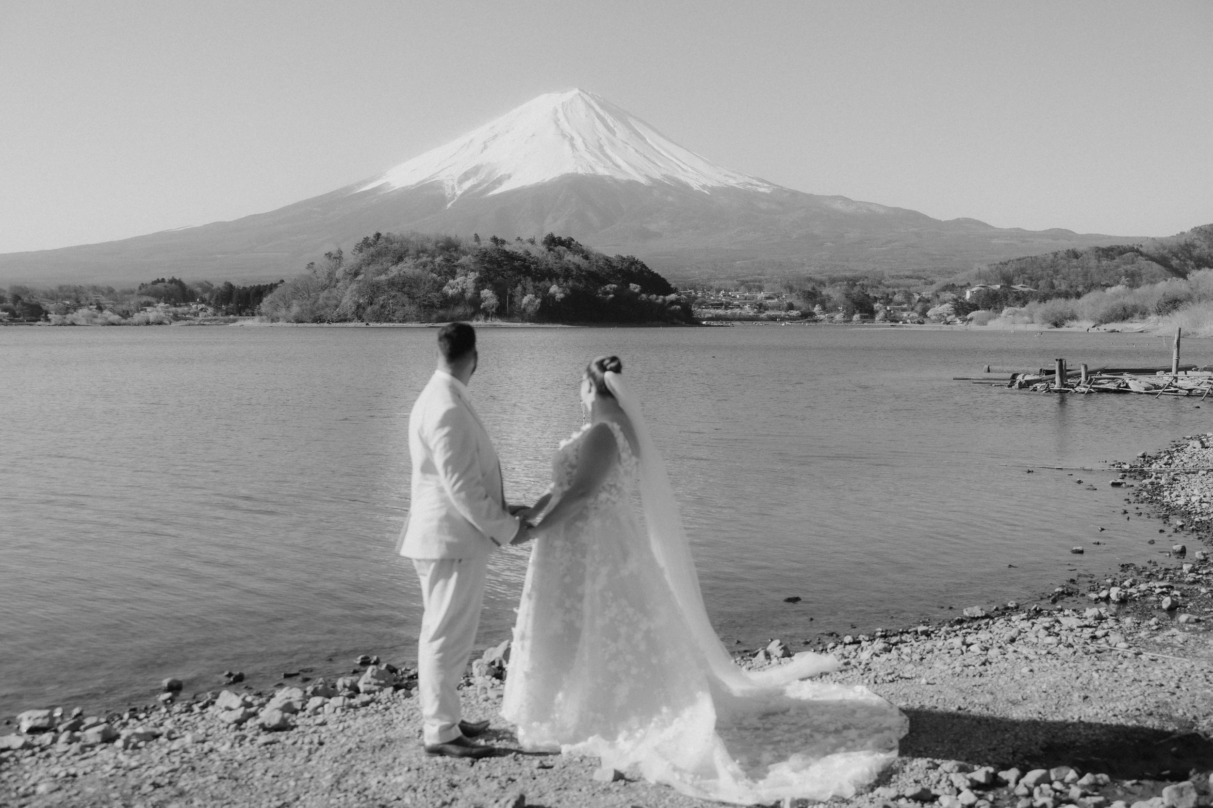 A couple in wedding attire holds hands while overlooking Mount Fuji and Lake Kawaguchi in black and white.