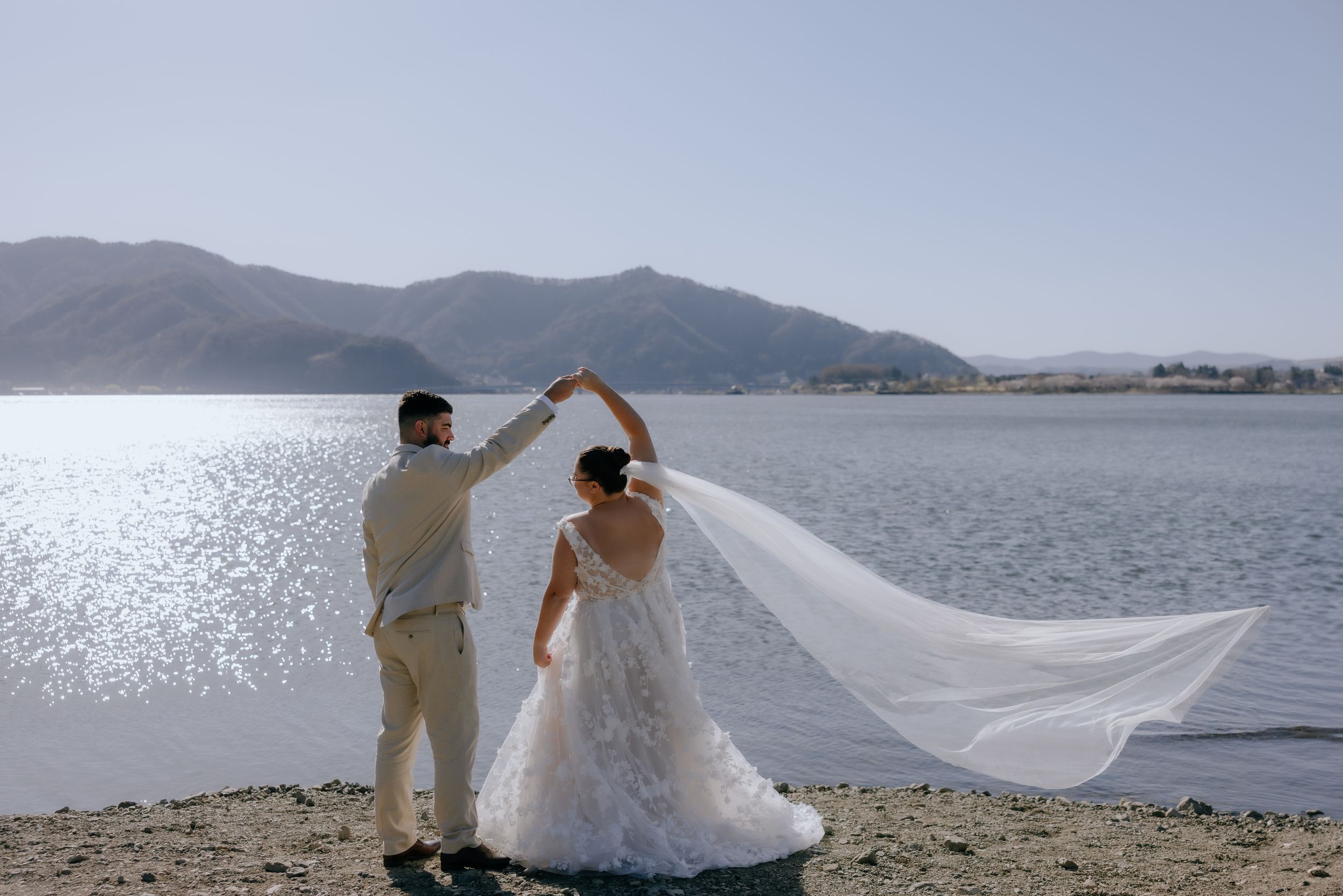 A couple in wedding attire dances together on a lakeside beach with mountains and sparkling water in the background.