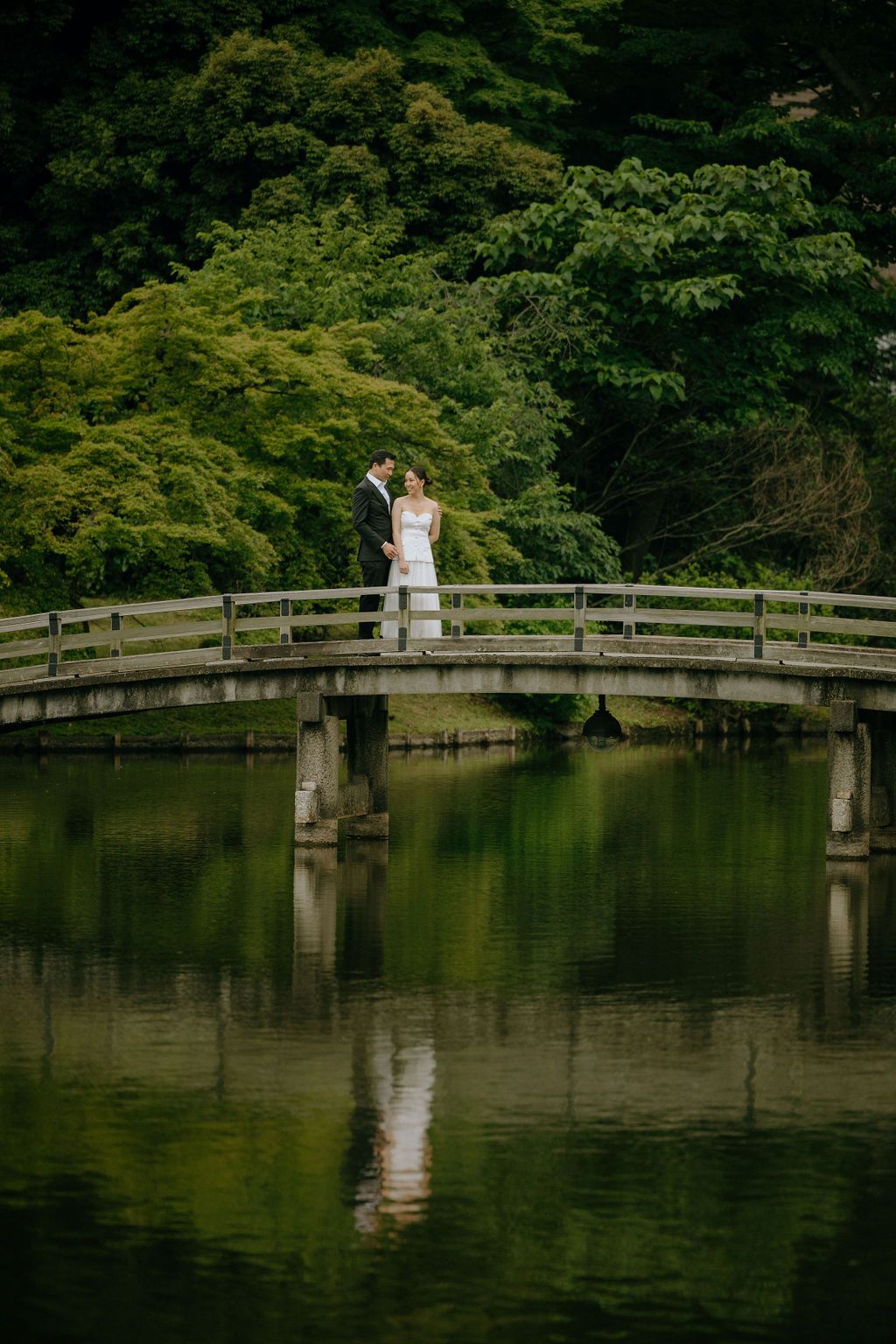 Wedding couple standing on a curved bridge over tranquil waters in a Japanese garden.