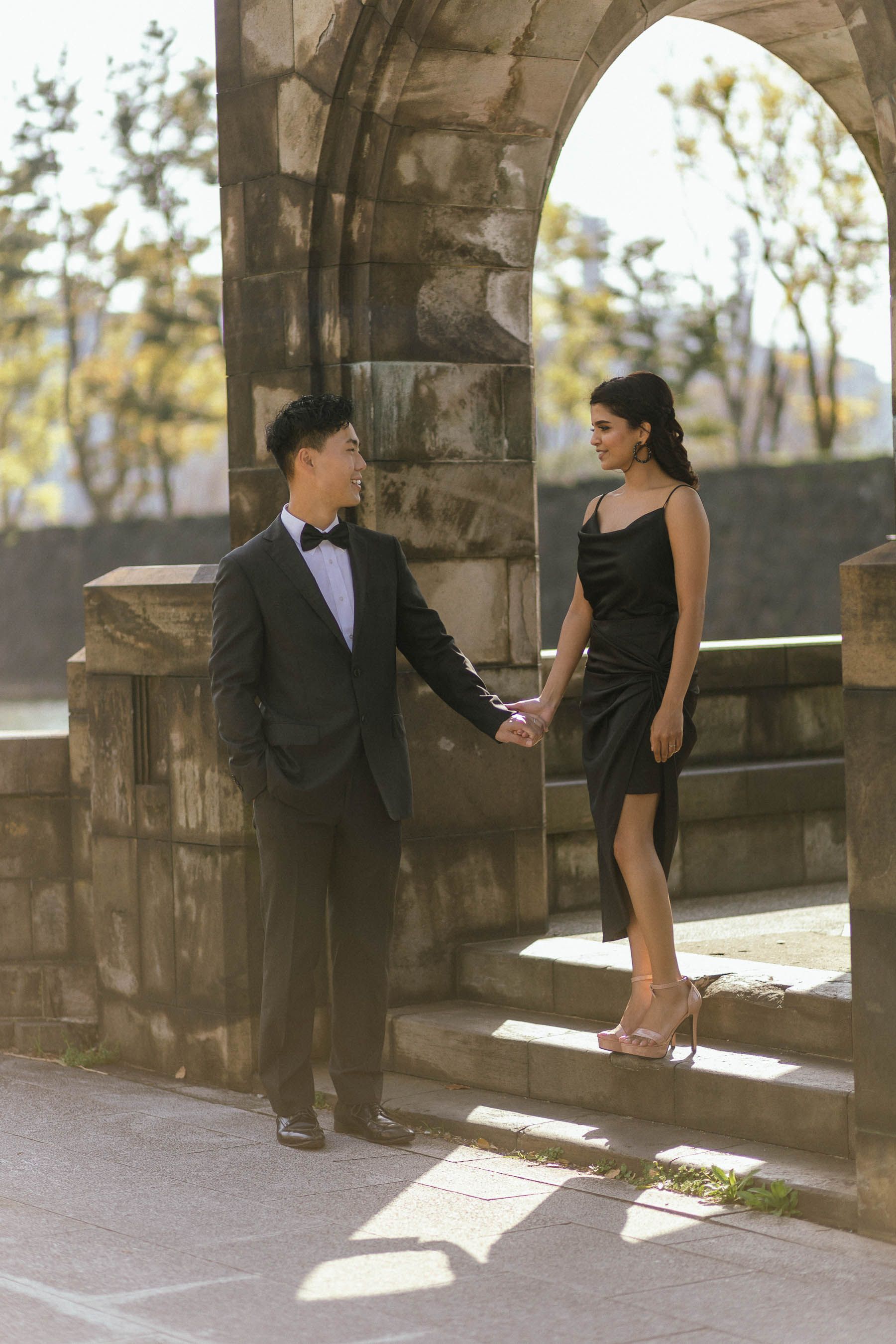 A couple in formal attire poses on stone steps under an ornate archway.