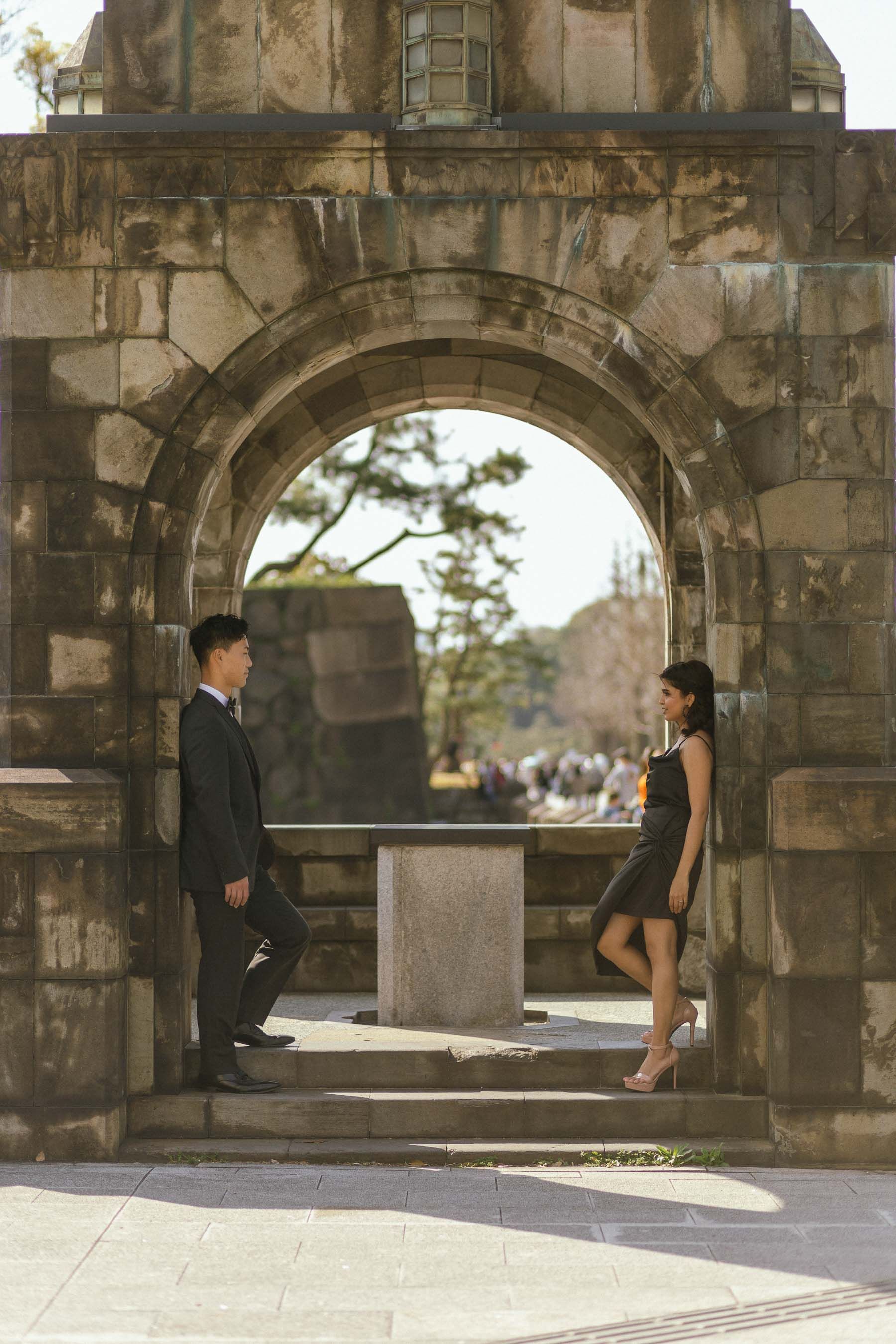 A couple in formal wear poses beneath a historic stone archway.