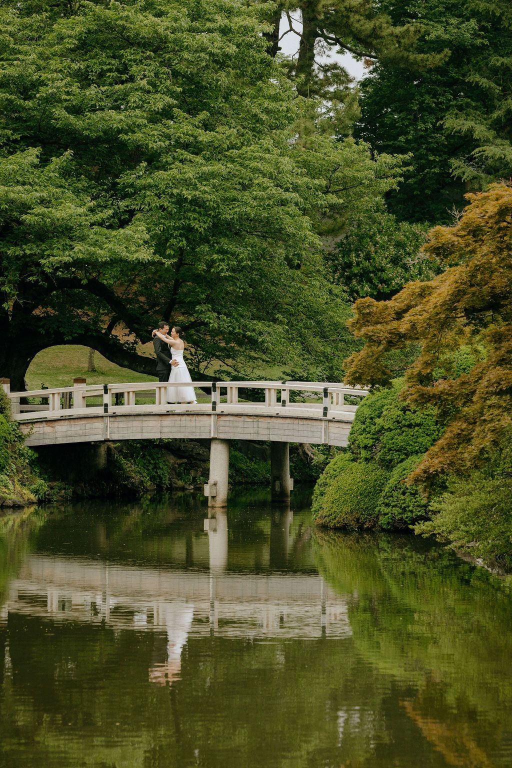 A curved wooden bridge reflects perfectly in still waters surrounded by Japanese maples and greenery.