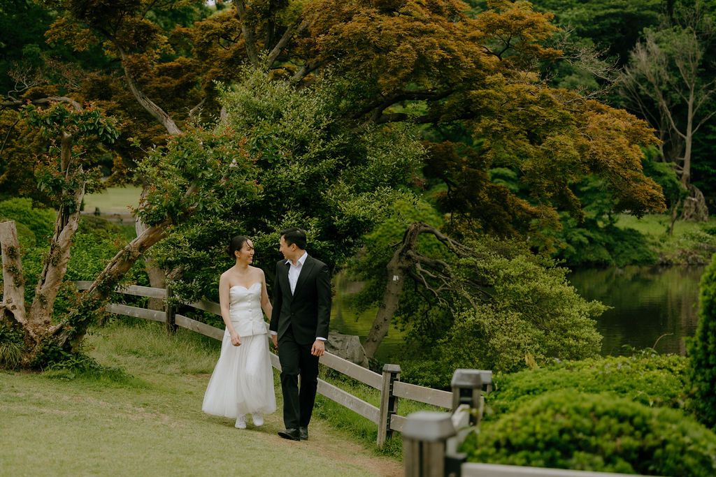 A couple strolls along a garden path with autumn foliage and a peaceful pond in the background.