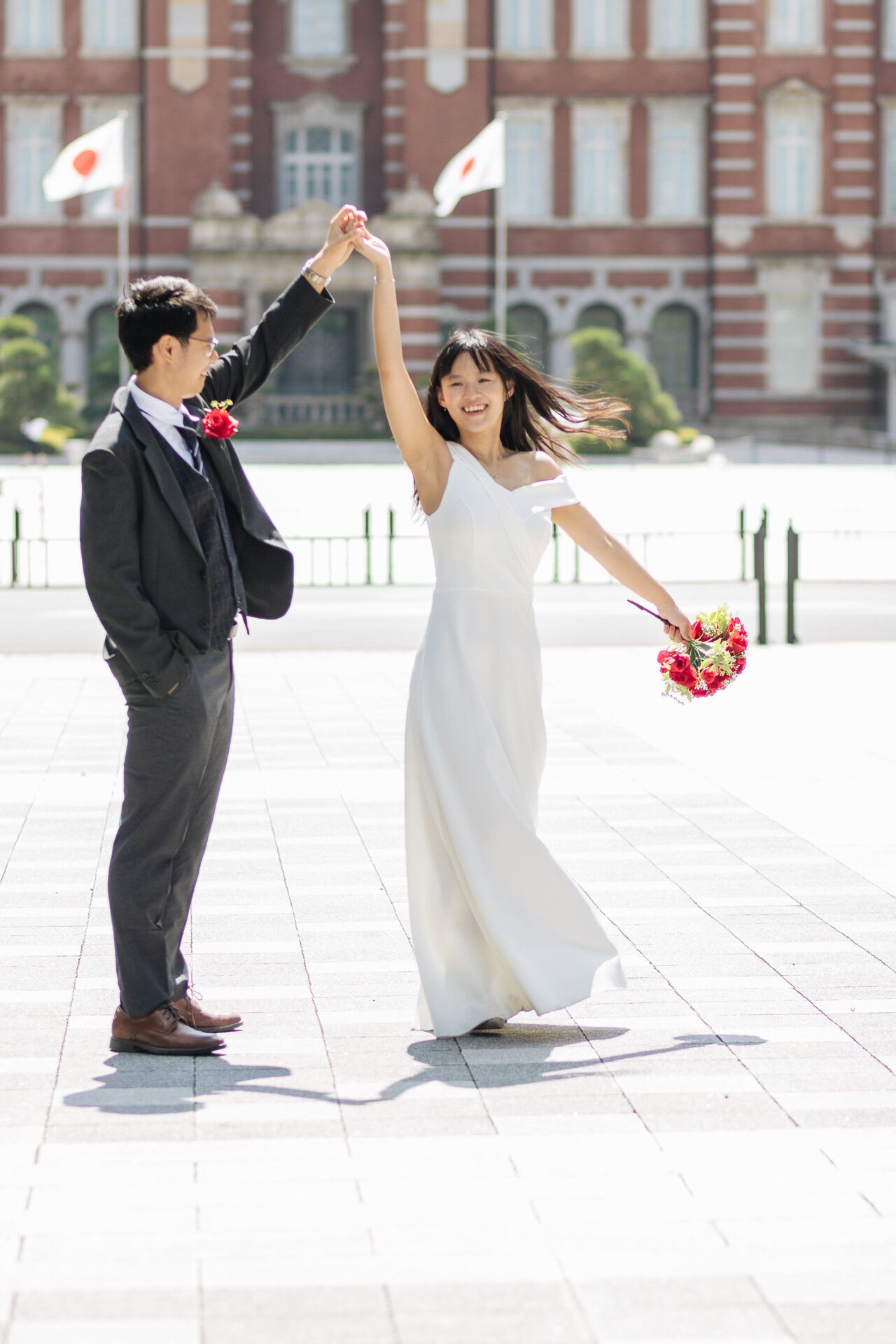 A couple dances and twirls with a red bouquet in front of Tokyo Station.