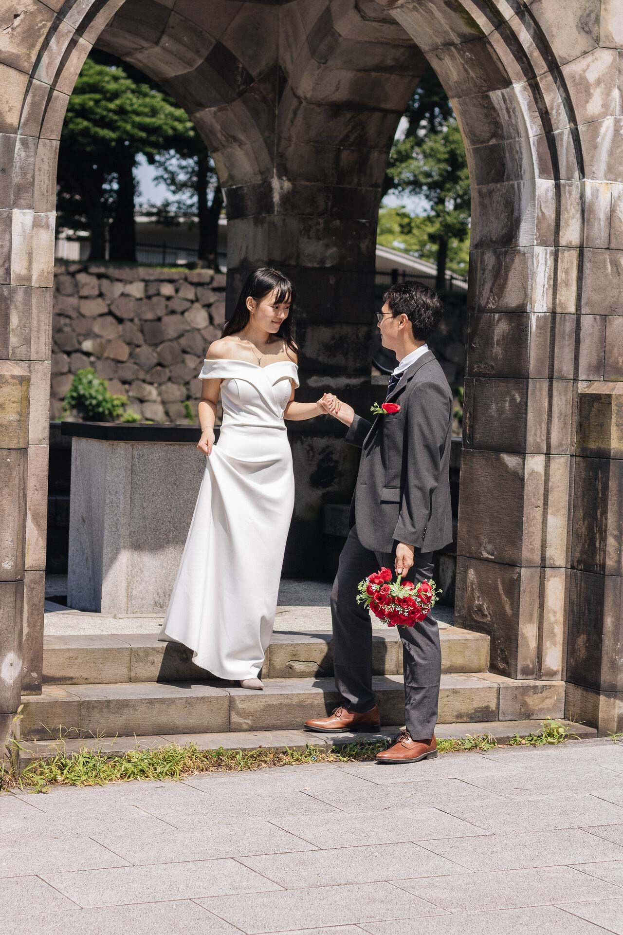 A couple stands in a stone archway during their wedding photoshoot with the bride in white and groom in gray suit.