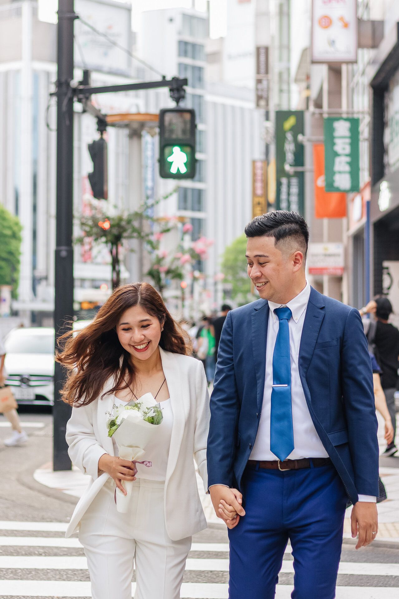 Stylish couple walking across city crosswalk in formal attire holding flowers.