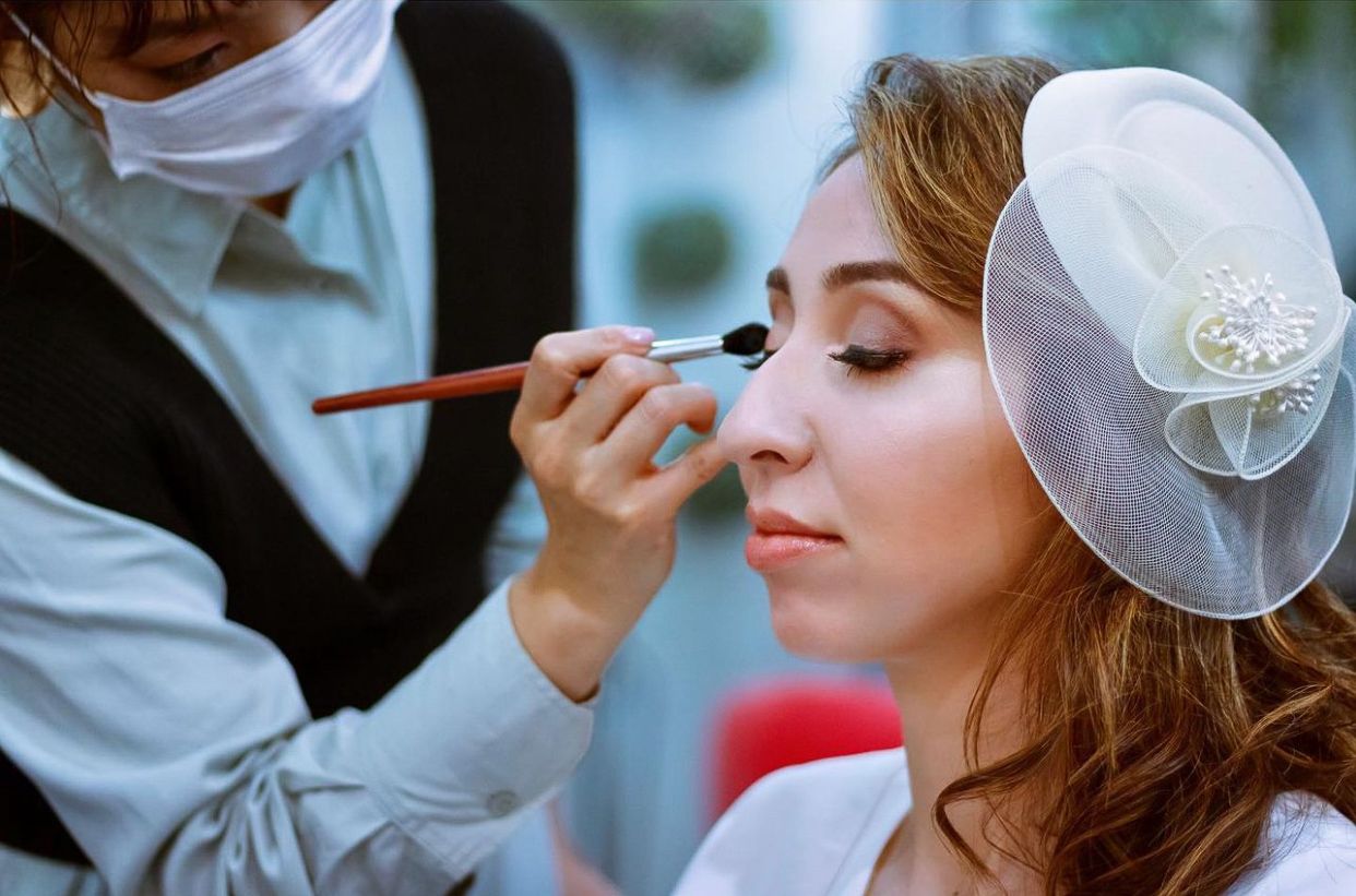 Professional makeup artist applying cosmetics to bride wearing delicate white headpiece.