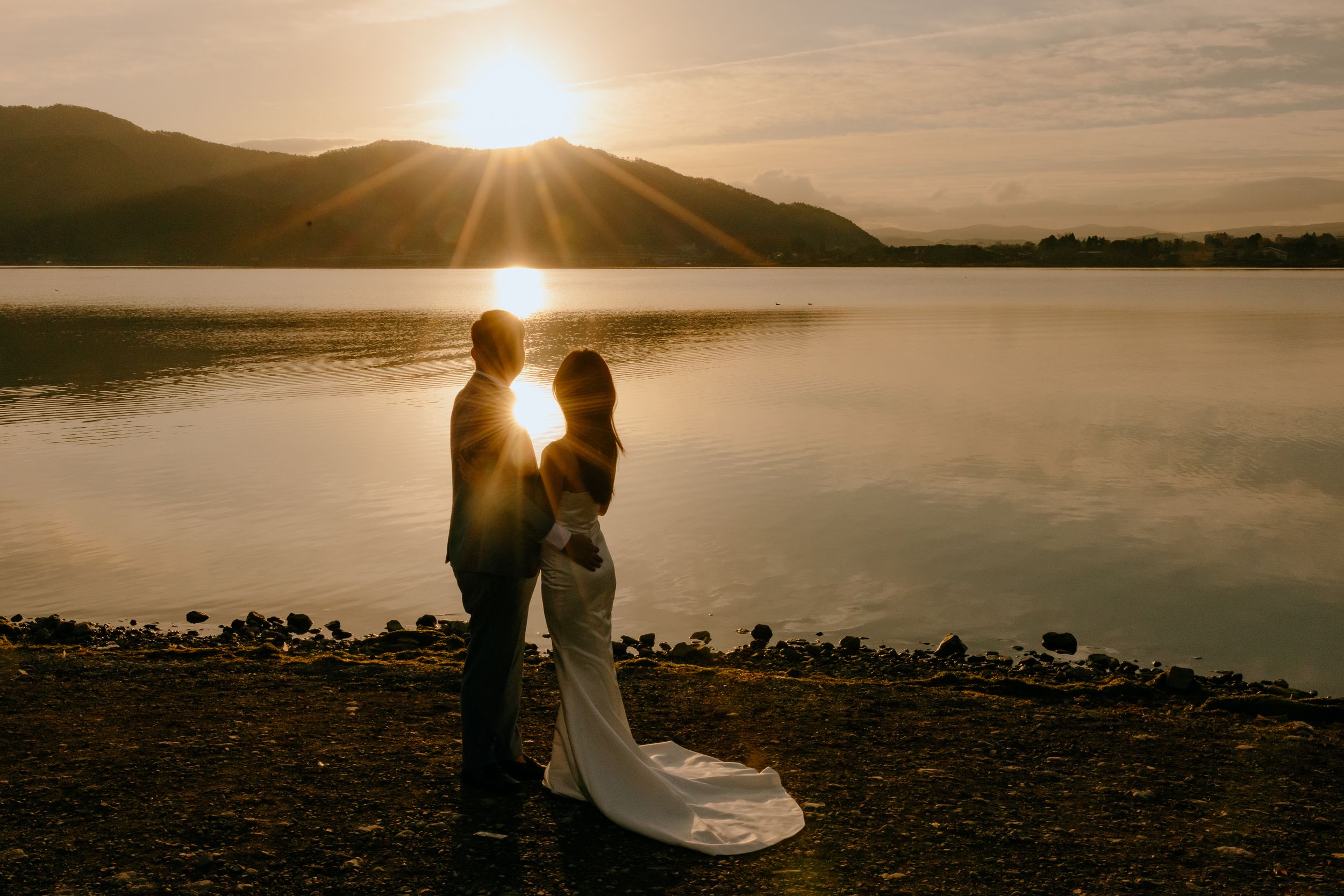 A couple embraces during sunset by a serene lake with dramatic golden light creating silhouettes against the water.