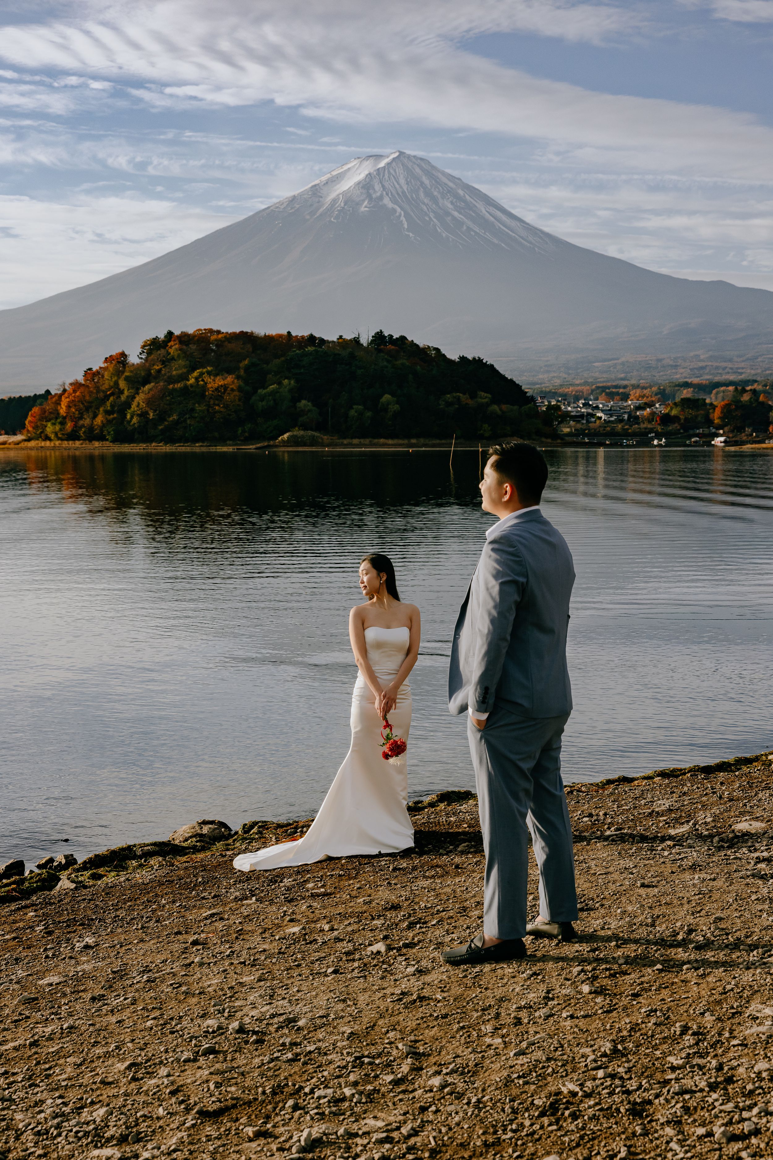 A wedding couple stands by the tranquil shores of Lake Kawaguchiko with majestic Mount Fuji rising in the background.