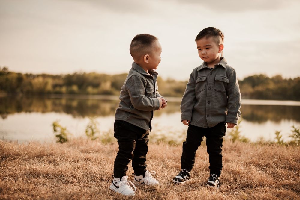 Two young boys in matching gray jackets and black pants stand facing each other on a grassy field near a lake.