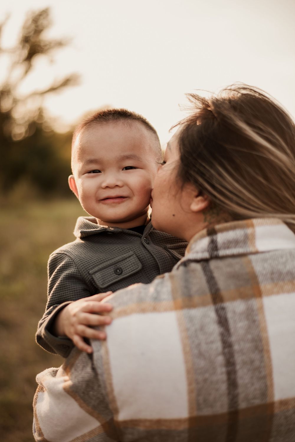 A smiling toddler boy being kissed on the cheek by his mother outdoors during golden hour sunset.