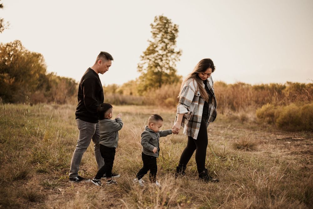 Family of four walking through golden autumn field at sunset, two young boys holding hands with parents in casual outdoor setting.