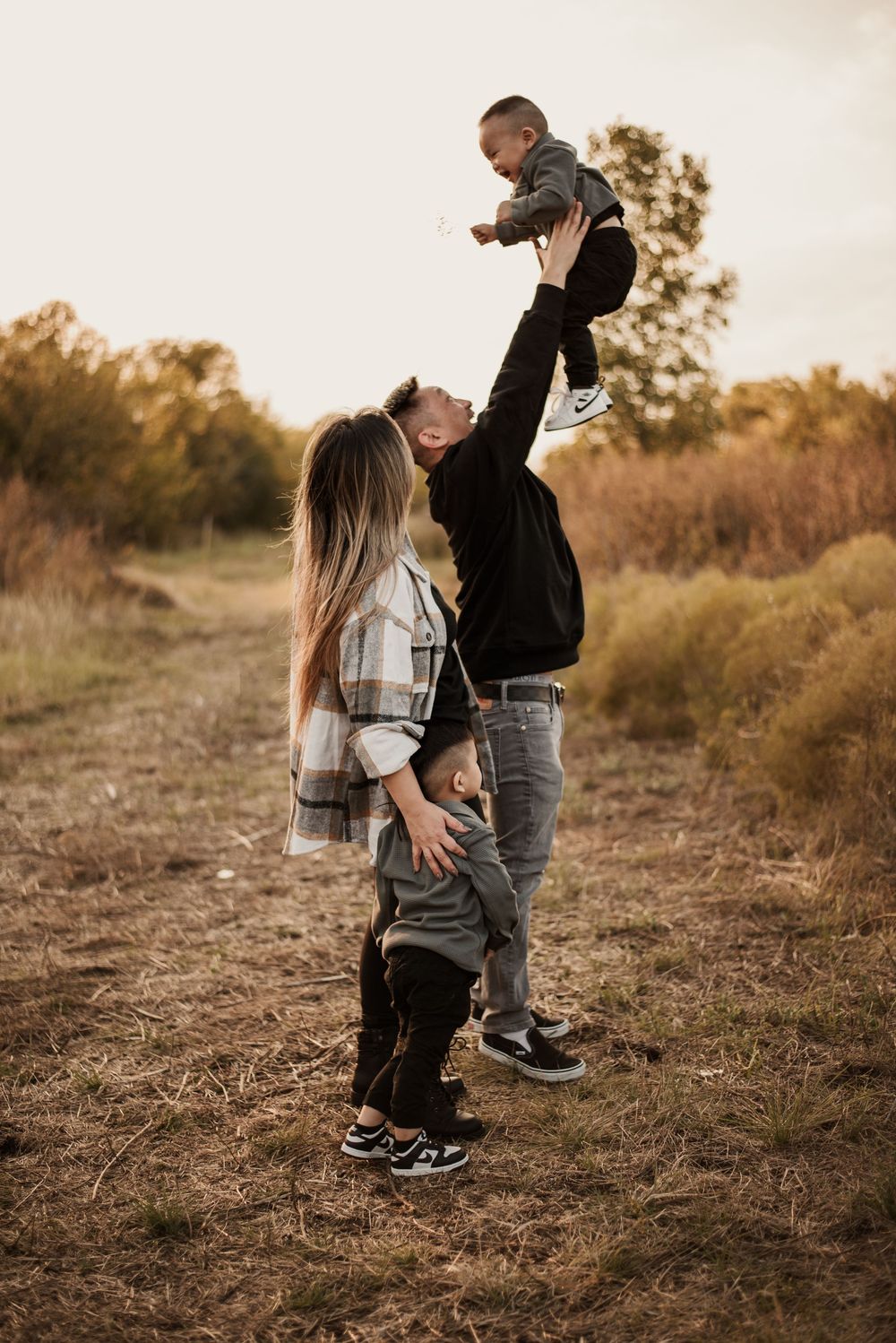 A couple plays with their two young children in a golden field at sunset, with the father lifting a baby in the air.
