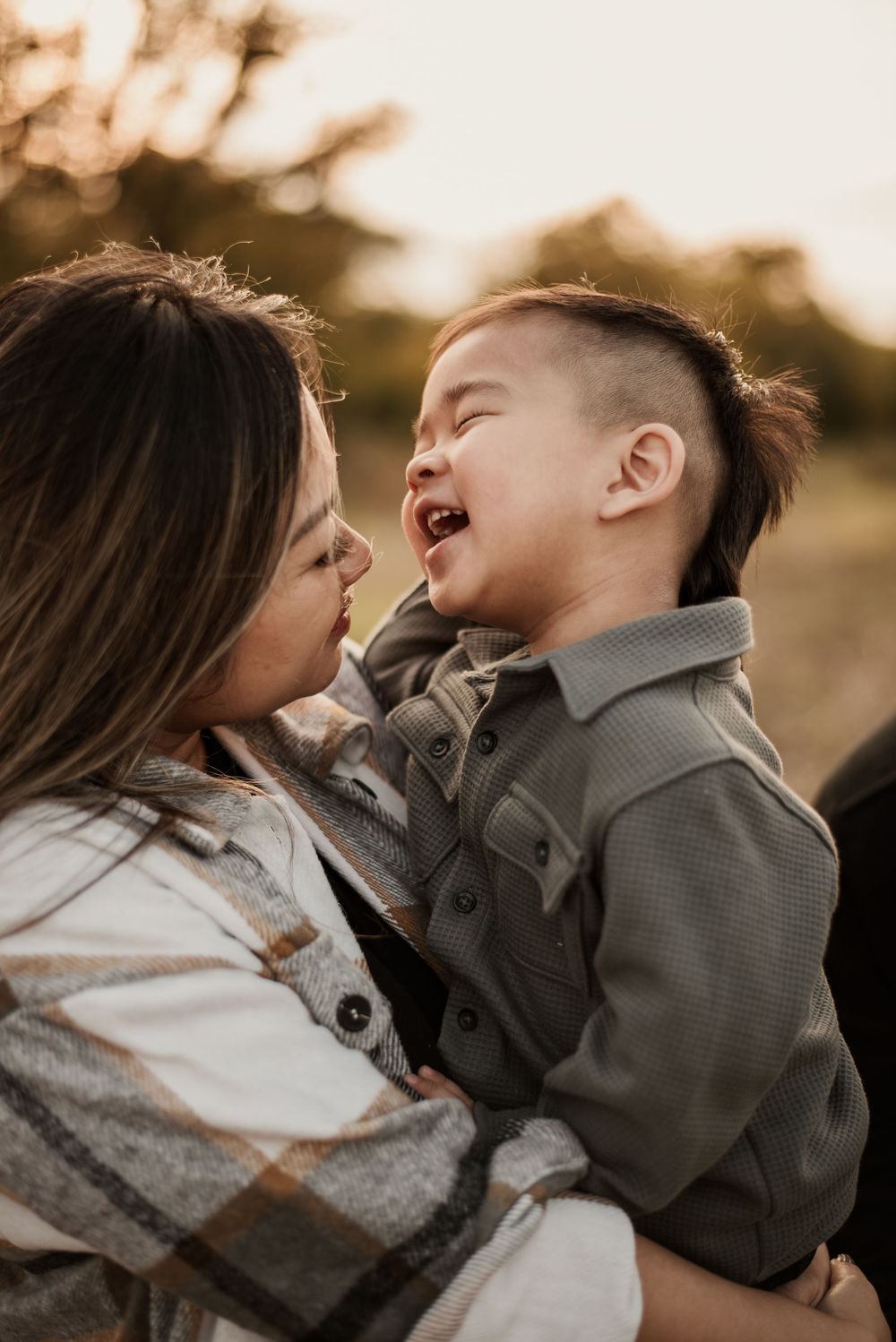 A joyful toddler laughs while being held by his smiling mother outdoors in warm golden sunset light.