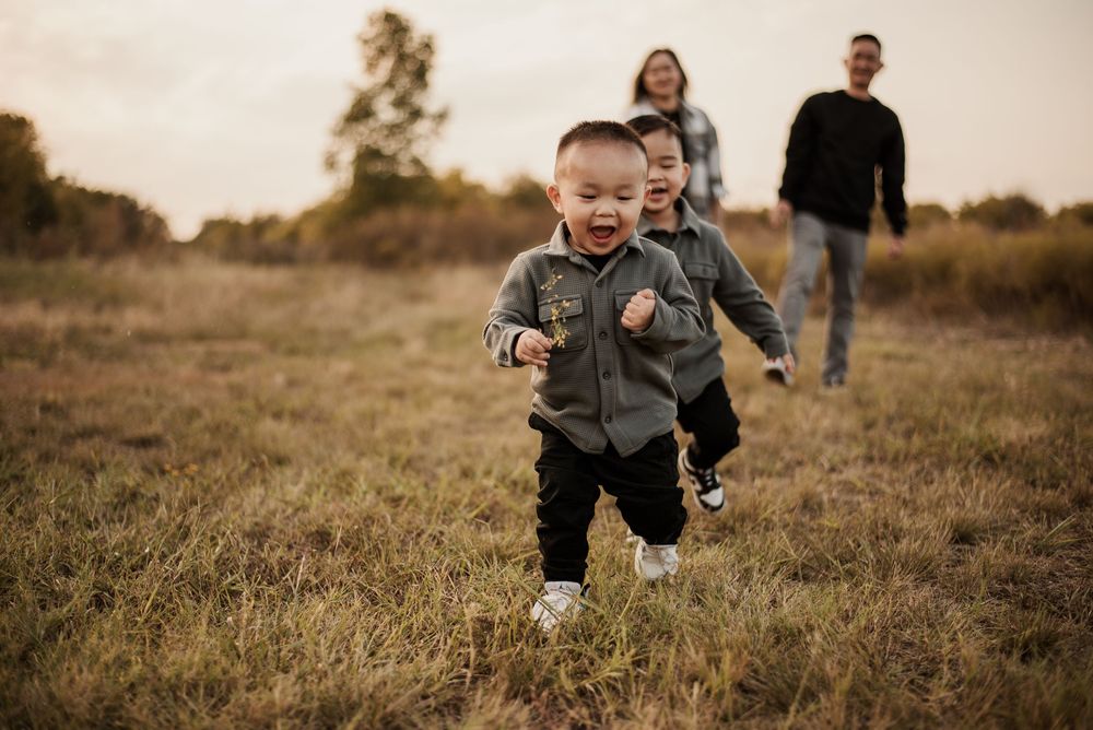 A joyful toddler runs through a grassy field while family members smile and follow behind during a golden hour outdoor photo session.