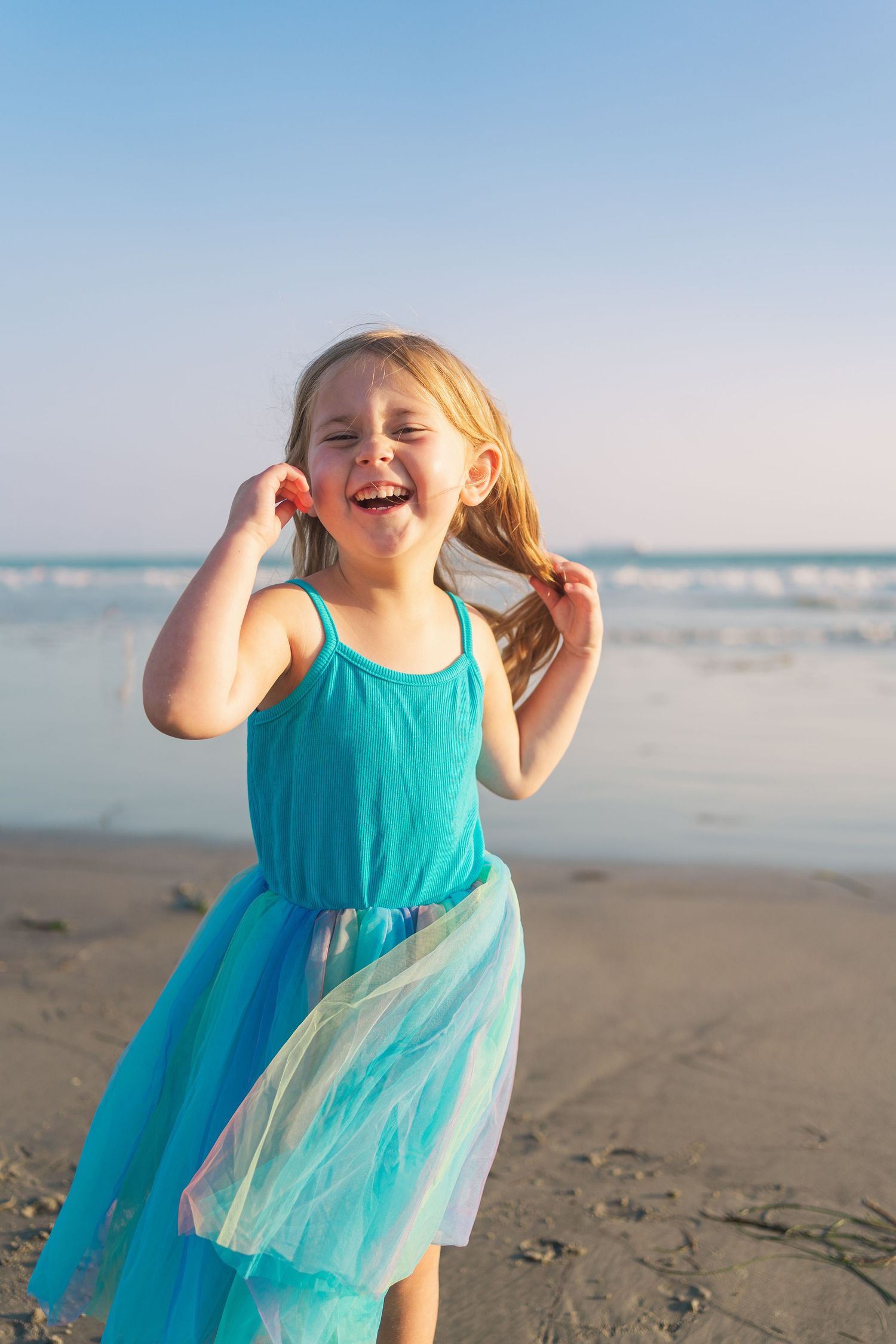 Child wearing turquoise dress plays joyfully on sunny beach with ocean waves in background.