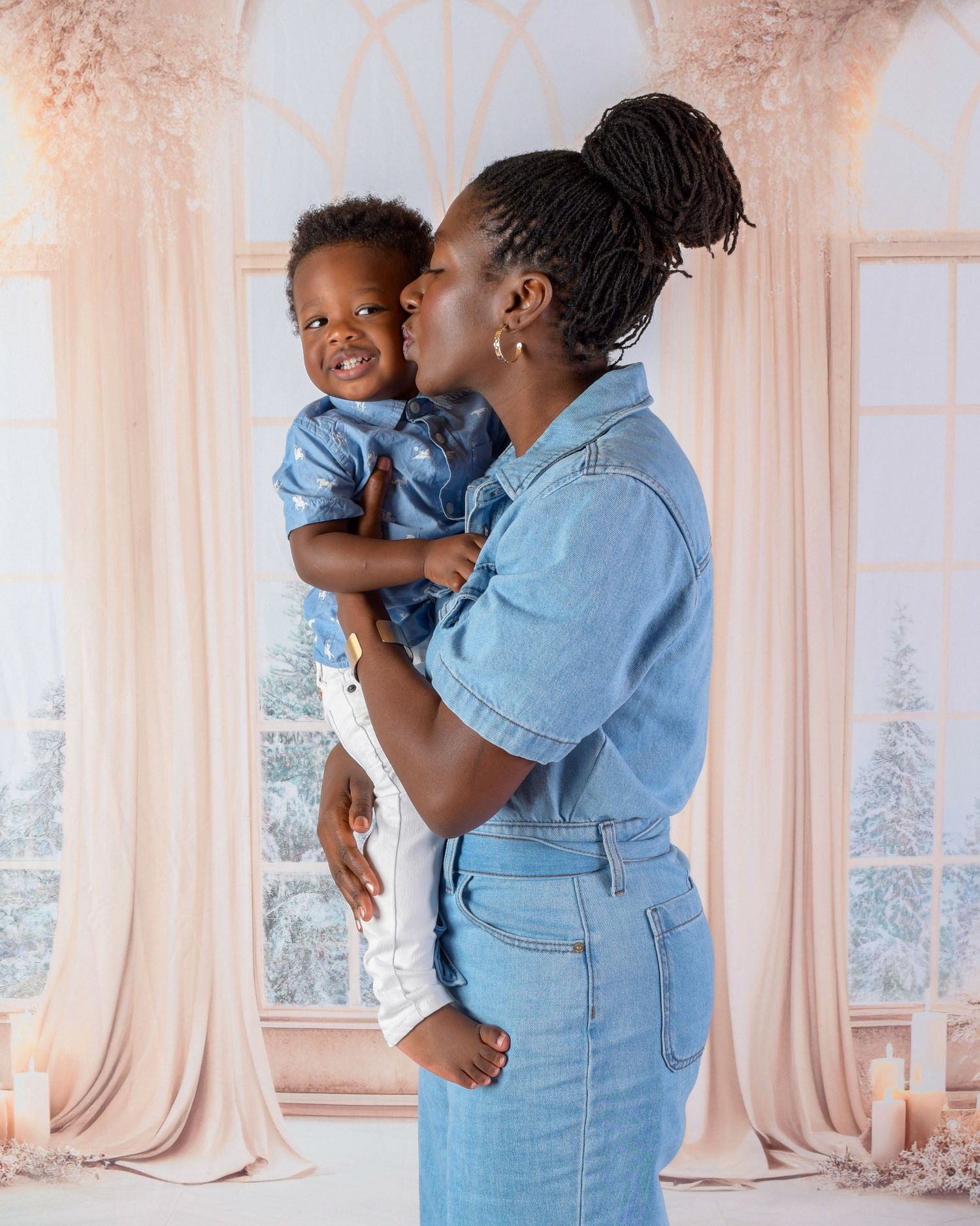 Parent in denim outfit giving affectionate kiss to toddler by window with snowy scene.