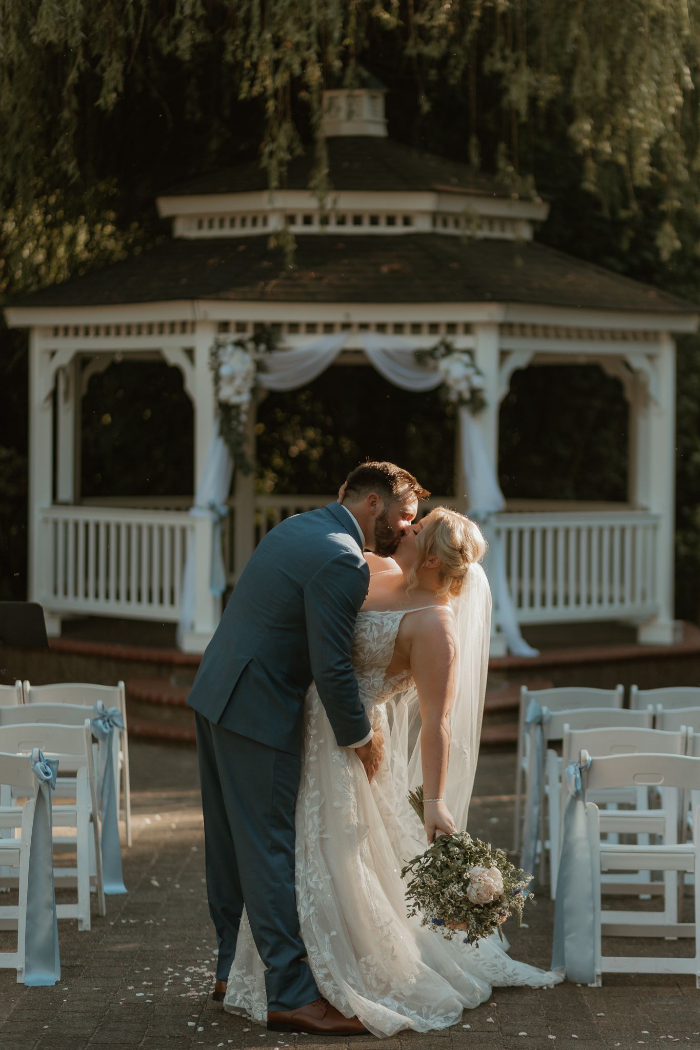 A romantic wedding couple sharing a kiss in front of a white gazebo with rows of chairs set up for the ceremony.