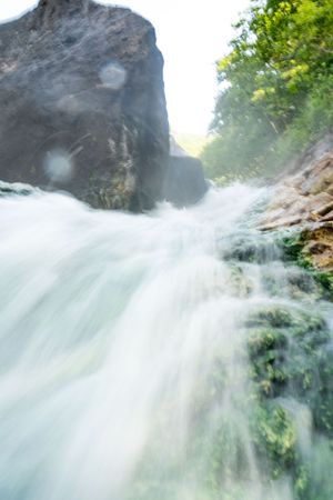 Misty waterfall flowing rapidly over rocks with sunlit vegetation in background.
