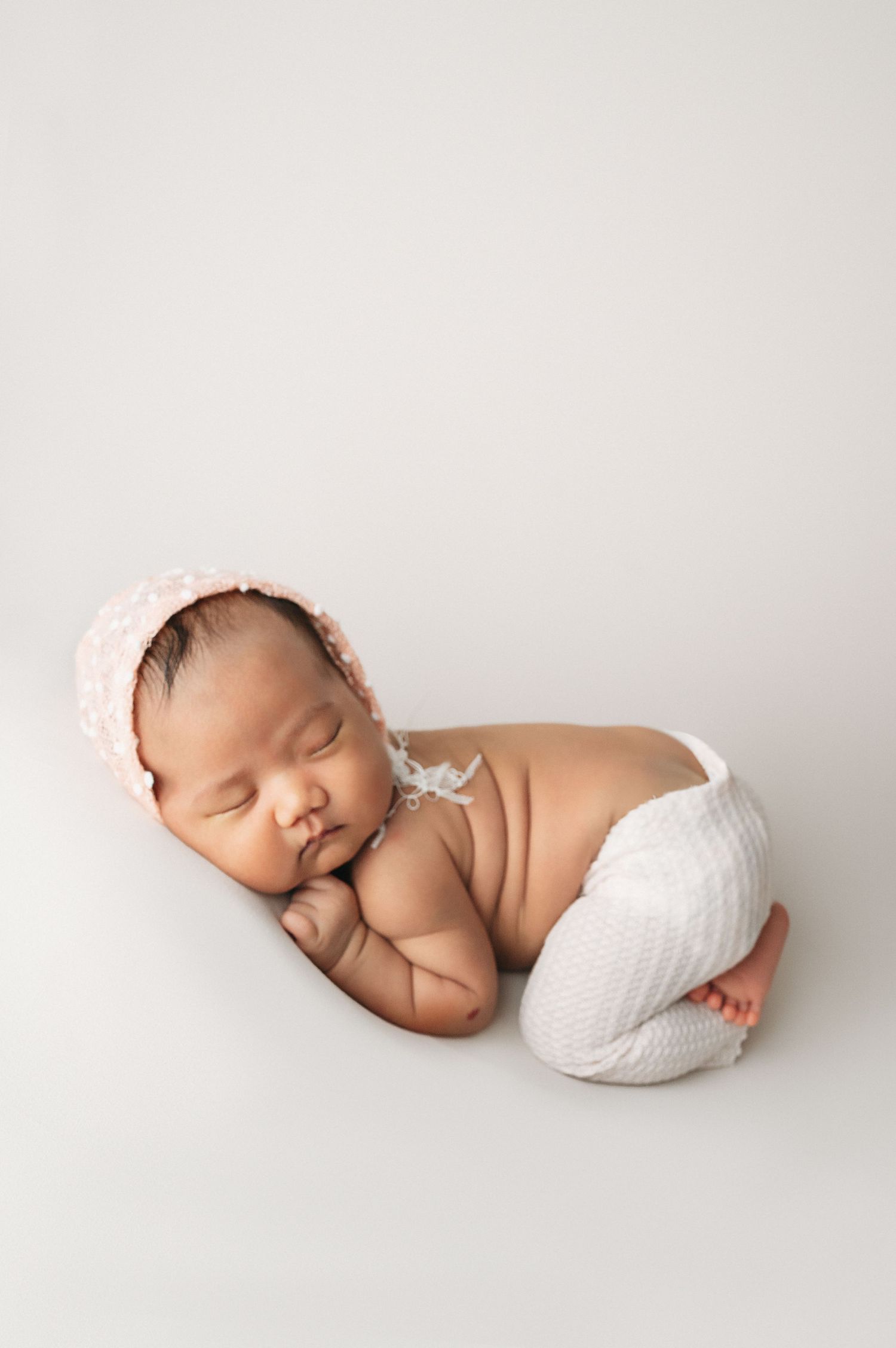 Sleeping newborn in white knit pants and pink bonnet posed on minimalist white background.