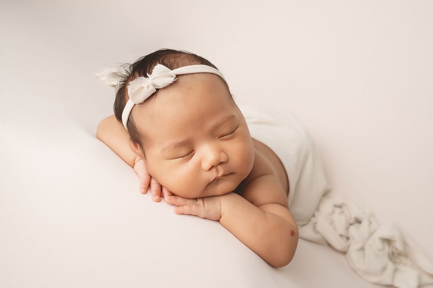 Newborn baby with white bow headband sleeping peacefully on white backdrop.