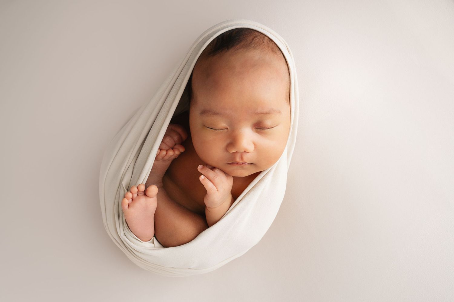 Sleeping newborn swaddled in white fabric against clean white studio background.
