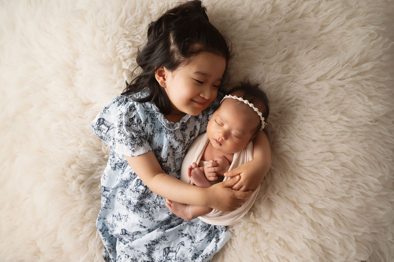 Siblings cuddling on plush white rug during newborn photo session.