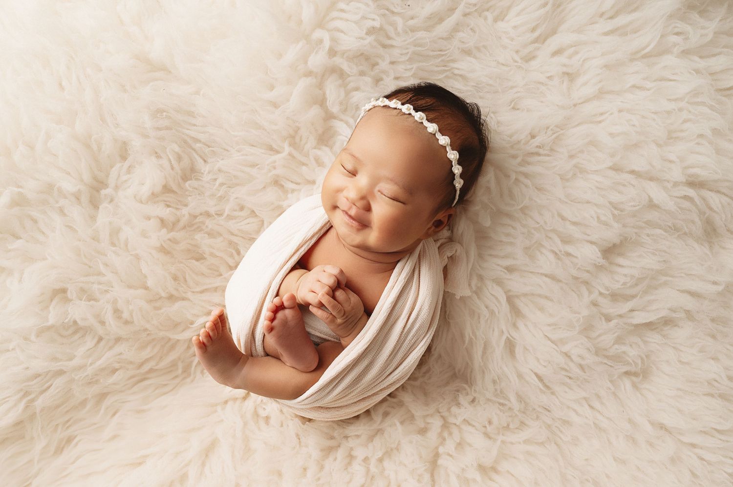 Newborn baby wrapped in white fabric sleeping peacefully on soft cream fur blanket with delicate headband.