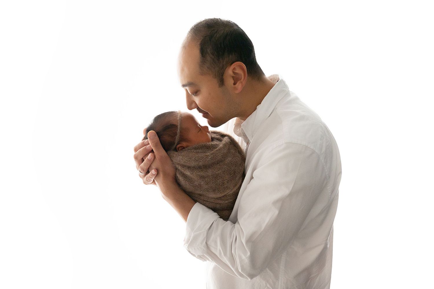 Intimate parent-child portrait with swaddled newborn against white backdrop.
