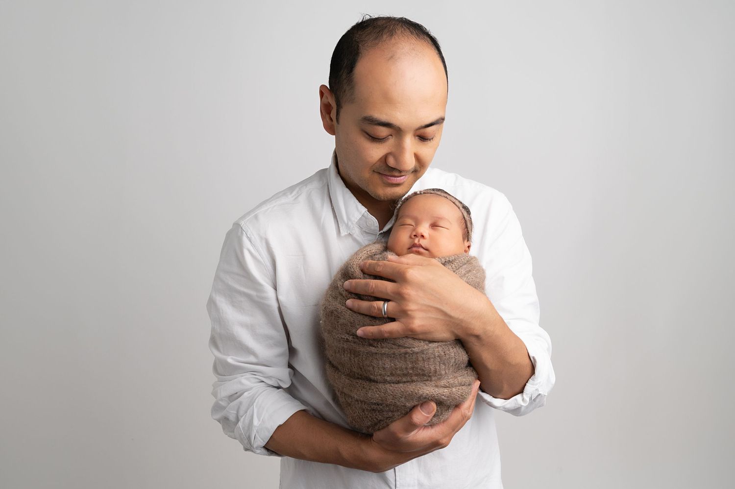 Parent in white shirt tenderly holding swaddled newborn against gray studio backdrop.