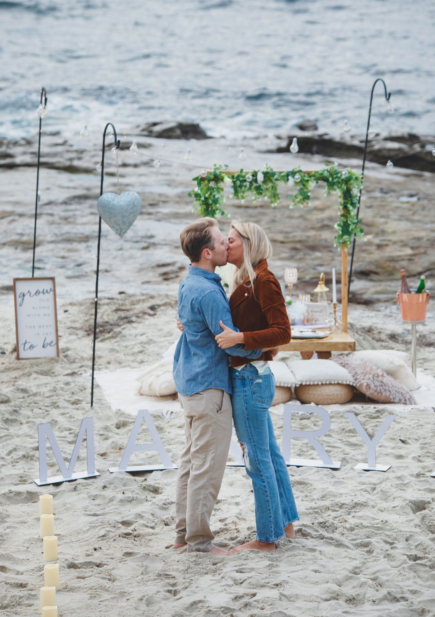 Romantic beach proposal setup with shepherd hooks and decorated arch on sandy shore.