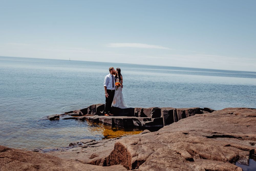 Wedding couple standing on rocky outcrop overlooking serene blue ocean waters.