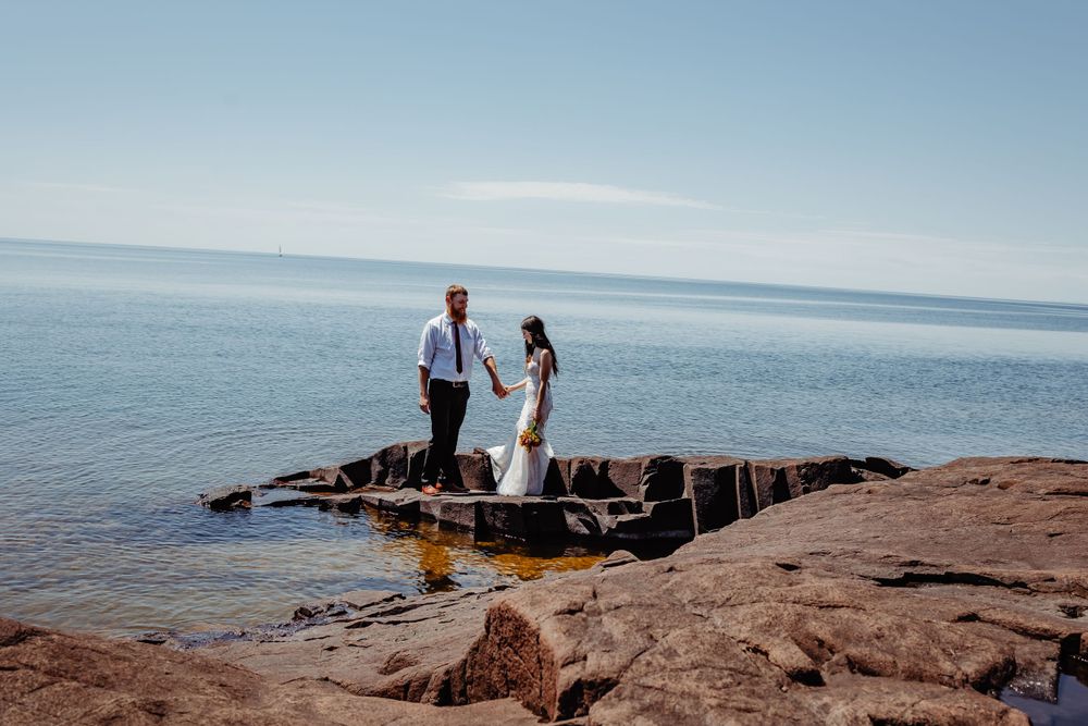 Couple in wedding attire standing together on rocky shoreline with ocean backdrop.