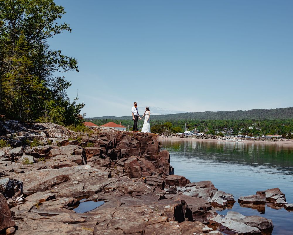 Two people celebrating on rocky cliffs overlooking a peaceful lake and forest landscape.