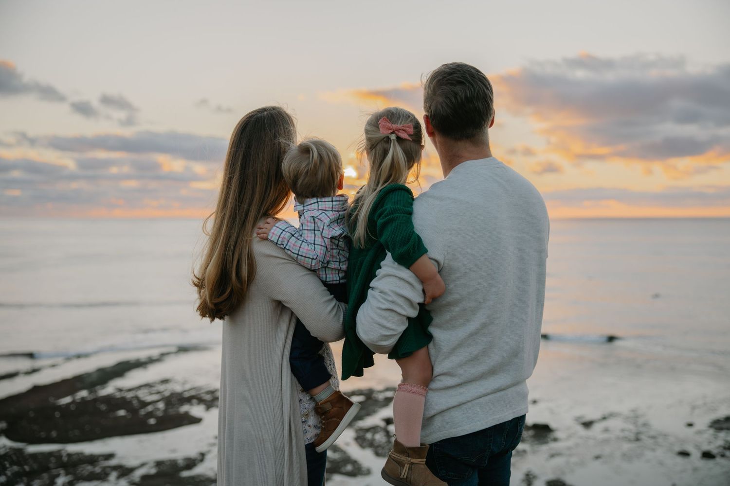 A family in matching grey outfits watches the sunset together by the ocean.