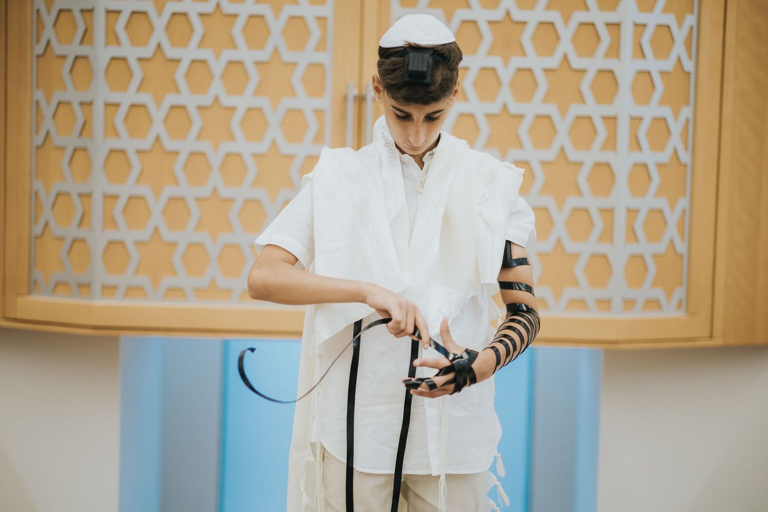 A person wearing a kippah and tallit reads from a prayer book against a geometric patterned wall.